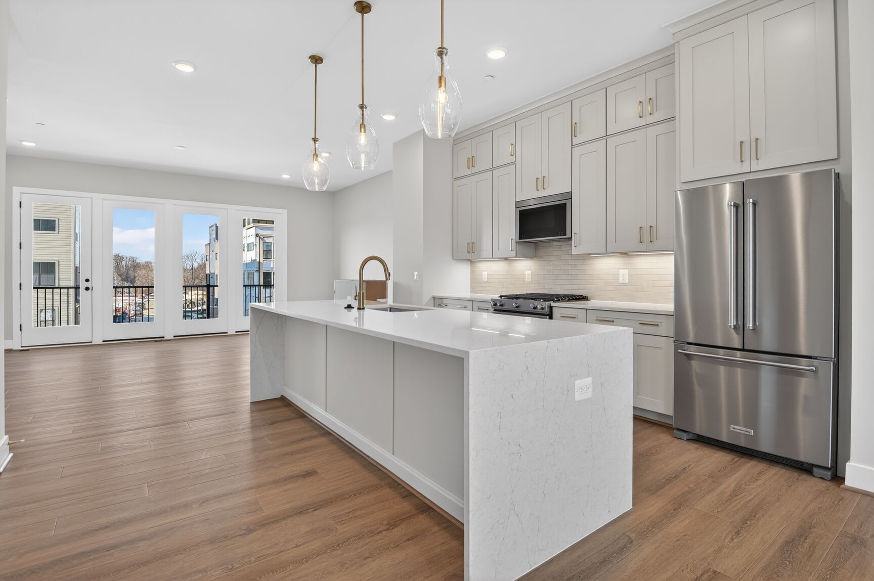 A modern, open-concept kitchen with white cabinets, stainless steel appliances, and a large island countertop, set against a backdrop of floor-to-ceiling windows offering a view of a cityscape.