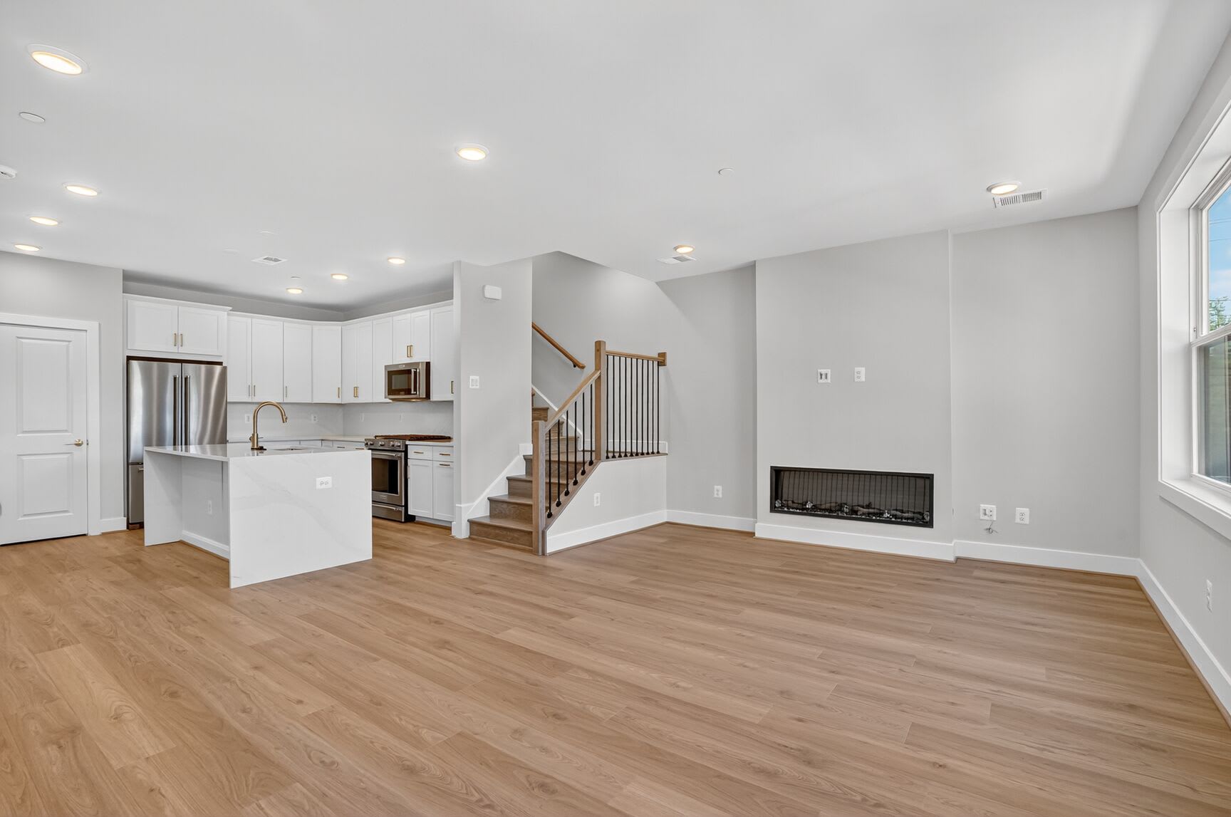 A spacious and modern open-concept living space with a kitchen, staircase, and a fireplace, featuring light-colored hardwood floors and neutral-toned walls.
