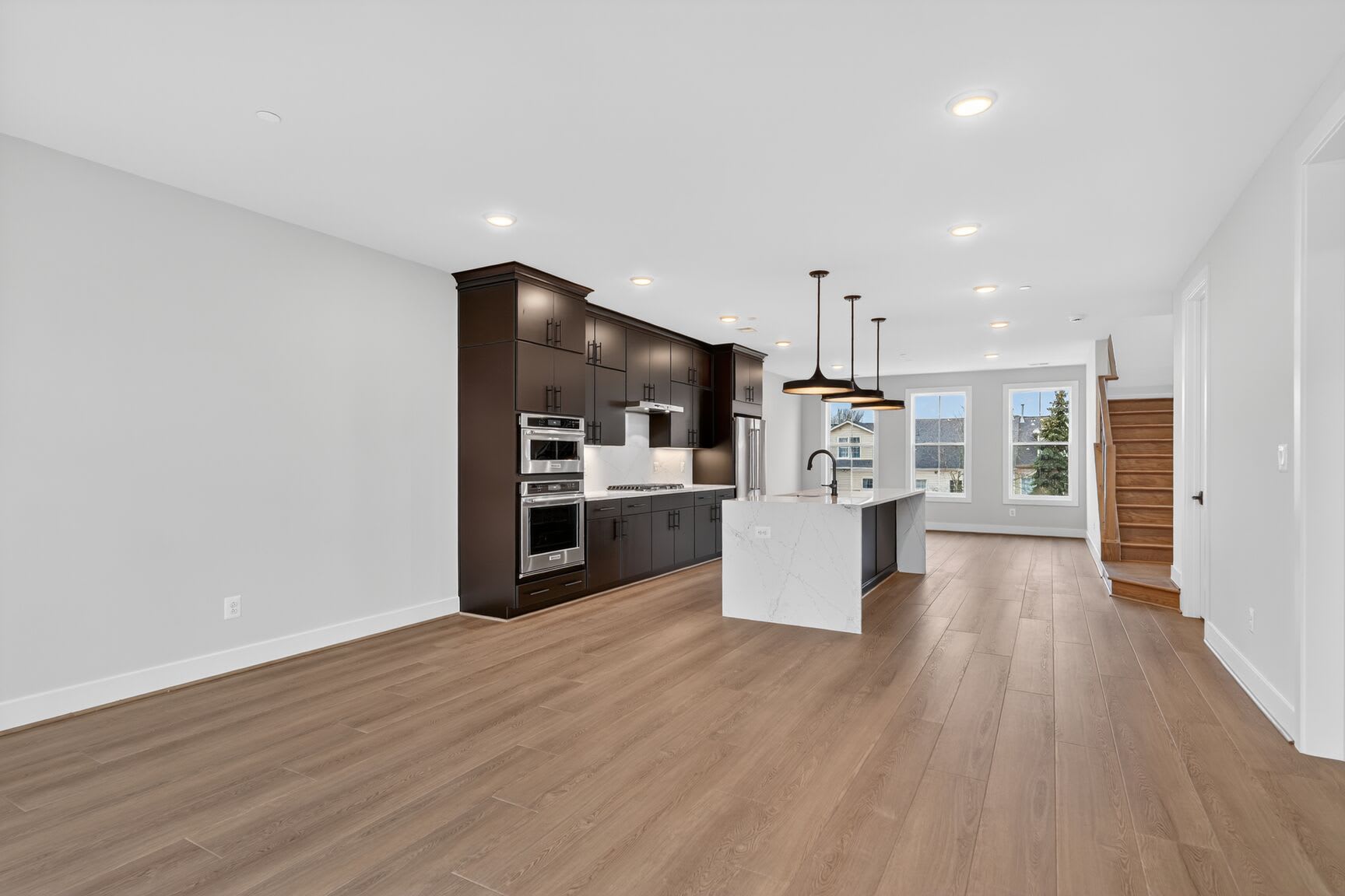 A modern, open-concept kitchen with dark wood cabinetry, stainless steel appliances, and a light-colored hardwood floor extends into a bright, airy living space.