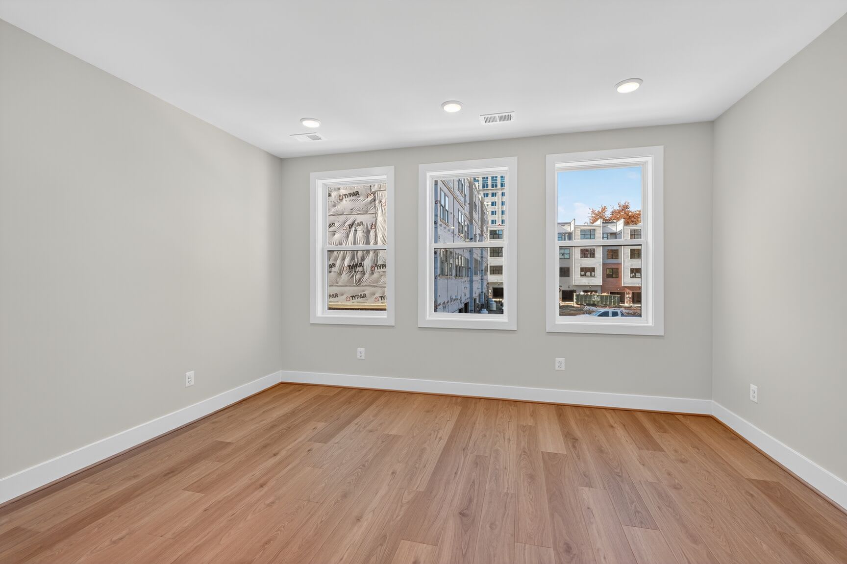 A spacious, well-lit room with large windows overlooking a cityscape, featuring hardwood floors and a neutral color scheme.