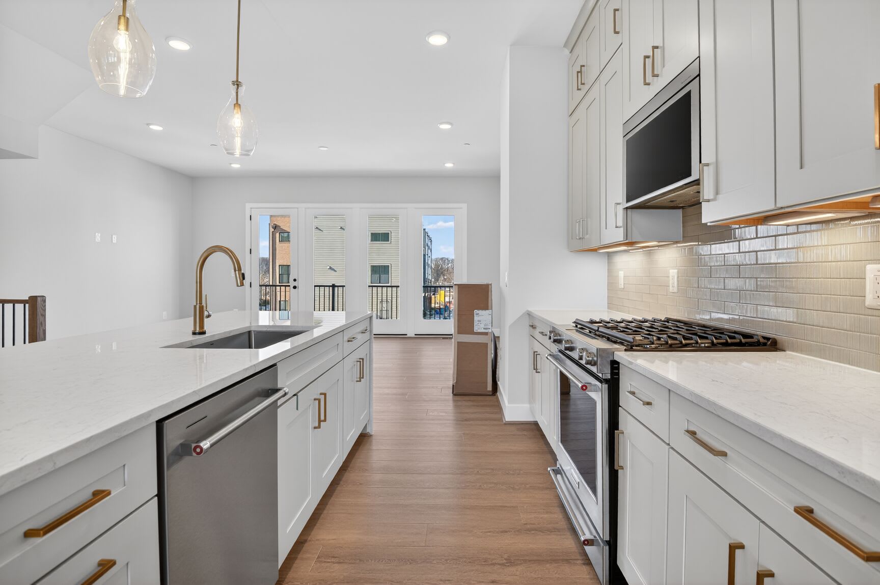 A modern, well-lit kitchen with white cabinets, stainless steel appliances, and a subway tile backsplash, leading to a hallway with a glass door.