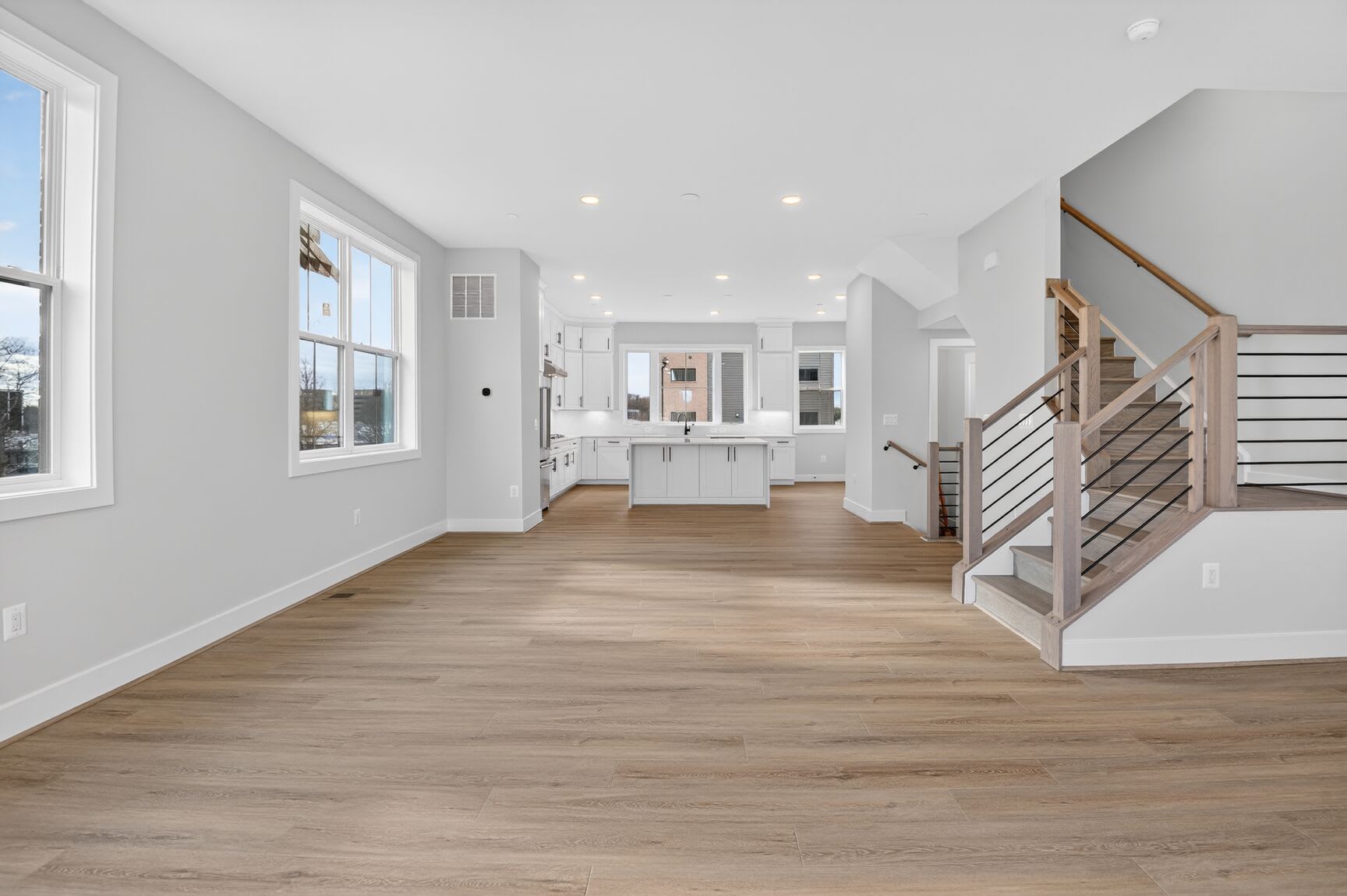 A spacious and bright open-concept living area with hardwood floors, a staircase, and a kitchen visible in the background.