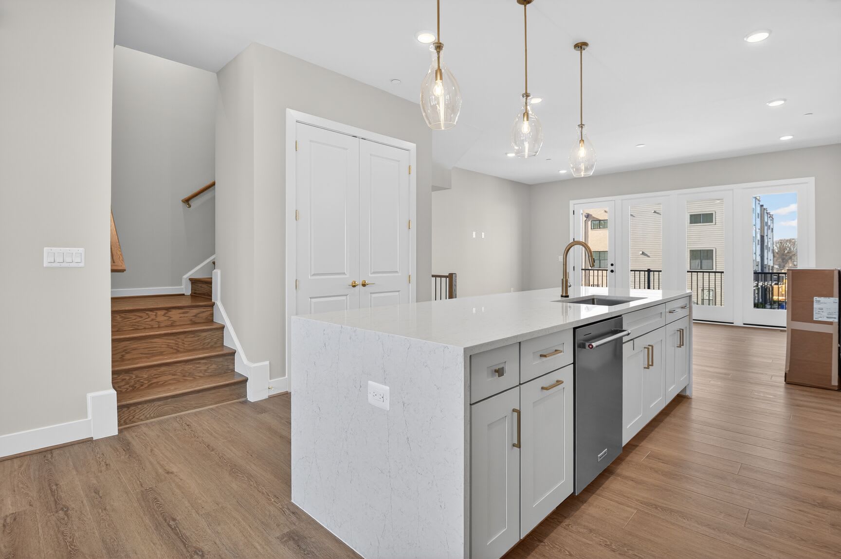 A modern, open-concept kitchen with white cabinets, a large island, and pendant lighting fixtures, set against a backdrop of hardwood floors and a staircase.