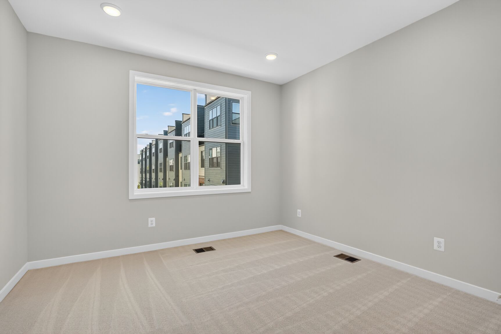 A spacious, well-lit room with a large window overlooking a modern urban landscape, featuring a hardwood floor and neutral-colored walls.