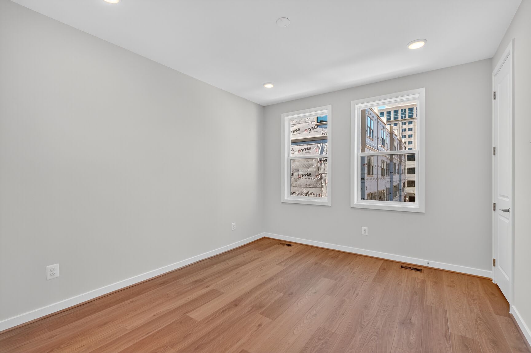 A spacious, well-lit room with light gray walls, hardwood floors, and large windows providing a view of the surrounding buildings.
