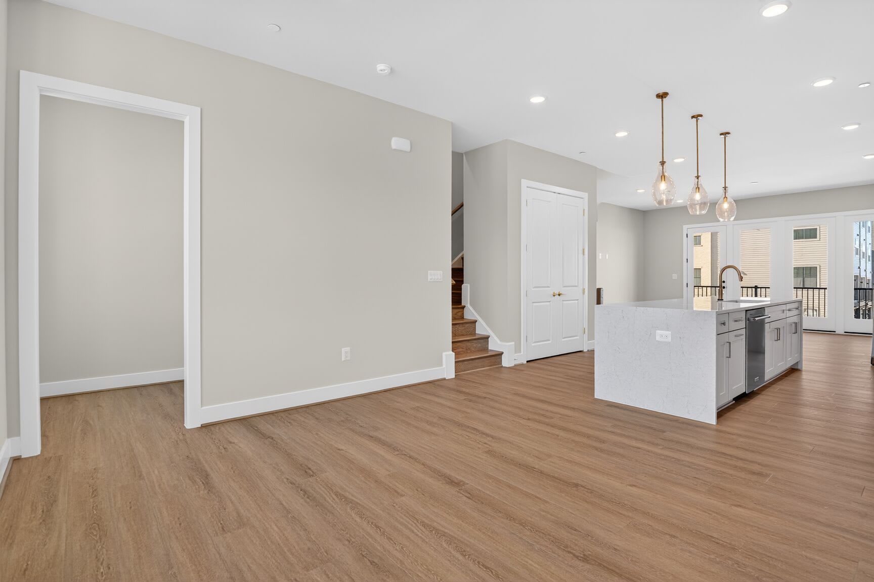A spacious and modern interior with light-colored walls, hardwood floors, and pendant lighting fixtures, leading to a kitchen area with white cabinets and appliances.