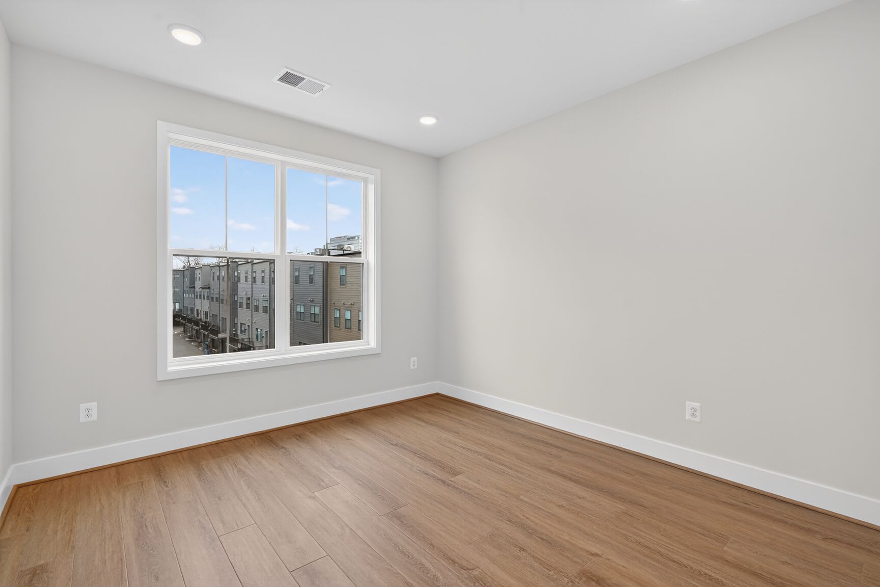 A spacious, well-lit room with a large window overlooking a cityscape, featuring hardwood floors and neutral-colored walls.