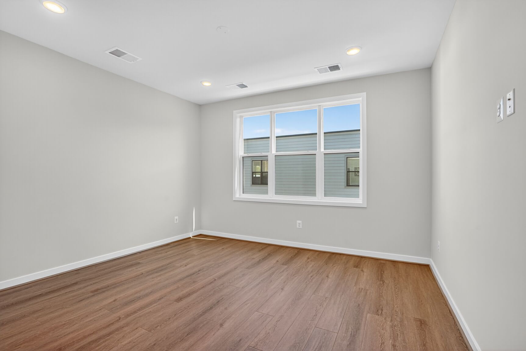A spacious, well-lit room with light gray walls, a large window, and a hardwood floor.