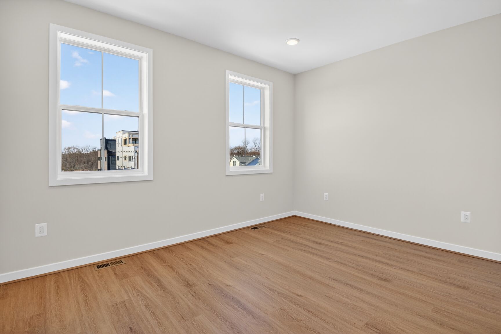 An empty room with large windows overlooking a residential neighborhood, featuring hardwood floors and neutral-colored walls.
