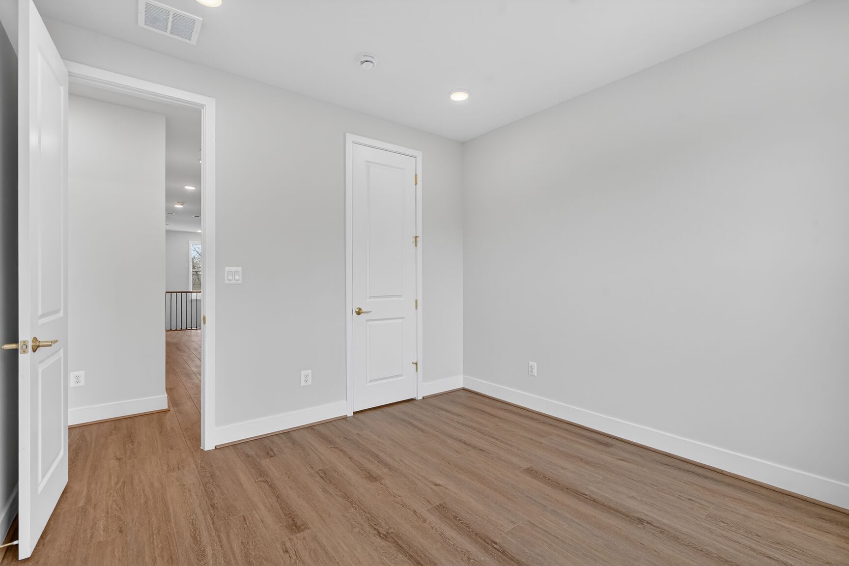 A spacious, minimalist room with white walls, a wooden floor, and a closed door leading to another area.