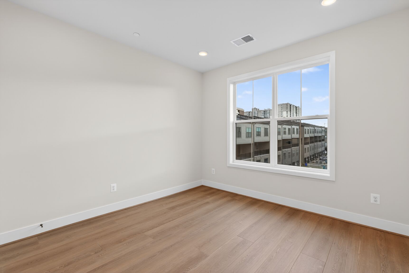 A spacious, well-lit room with large windows overlooking a cityscape in the background, featuring hardwood floors and a clean, minimalist design.