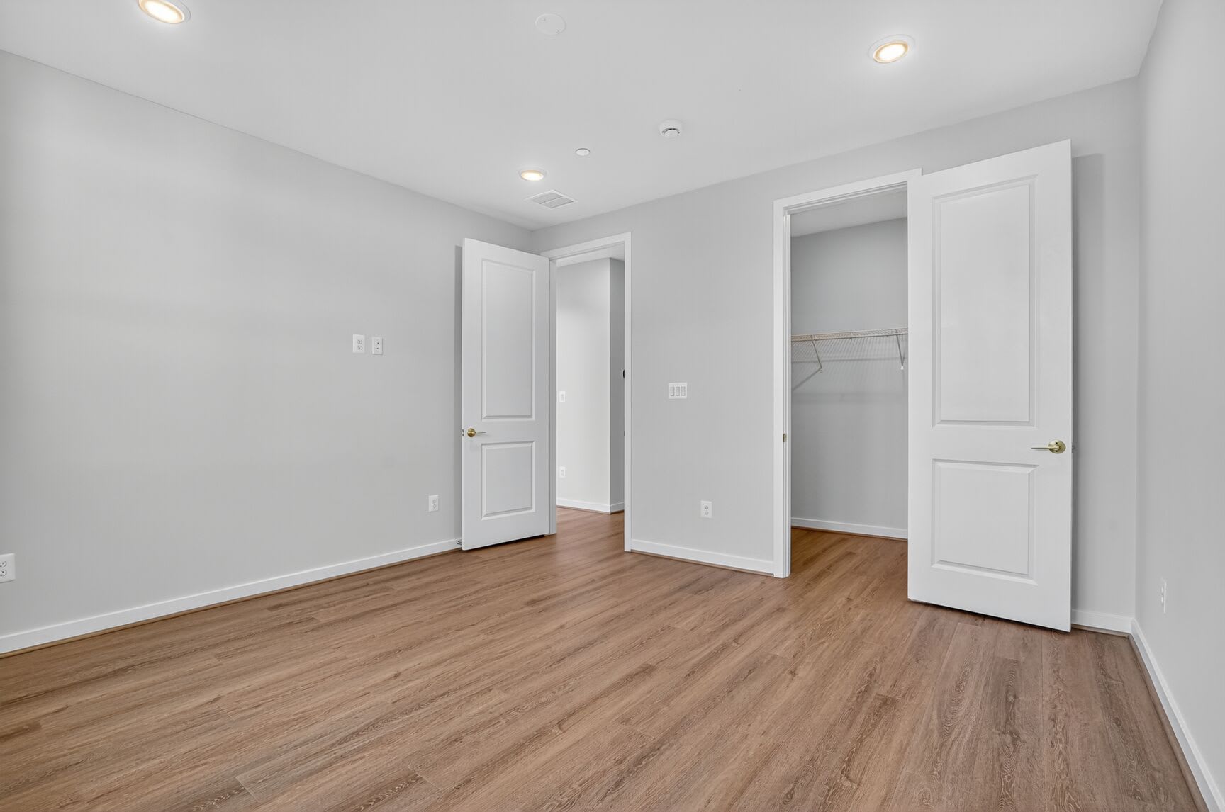 A spacious, minimalist bedroom with white walls, wooden flooring, and two closed doors, one of which appears to be a closet.