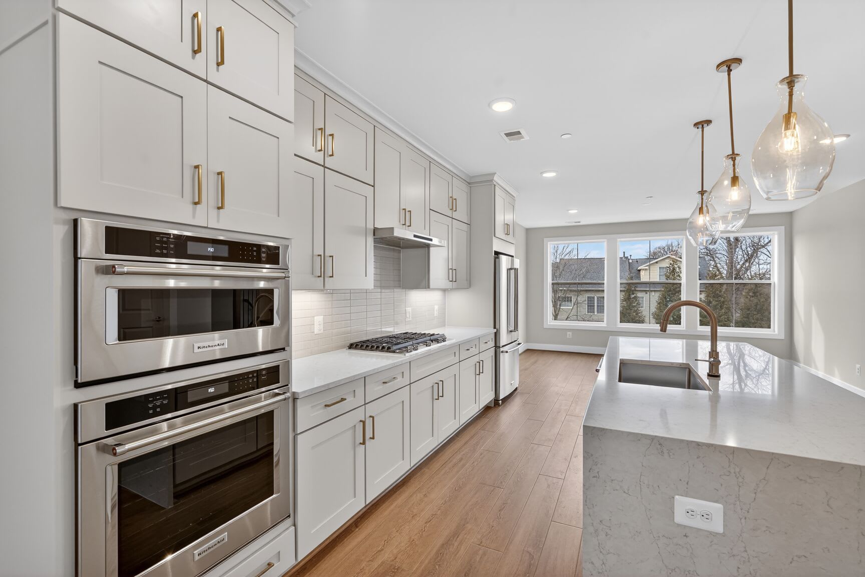 A modern, well-lit kitchen with white cabinets, stainless steel appliances, and a hardwood floor leading to a bright, open living space.
