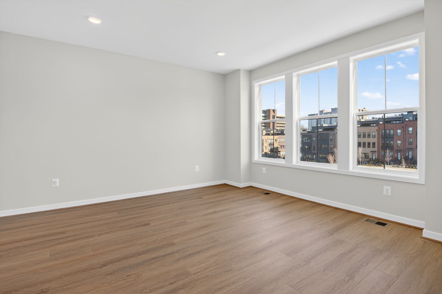 A spacious, well-lit room with large windows overlooking a cityscape, featuring hardwood floors and neutral-colored walls.