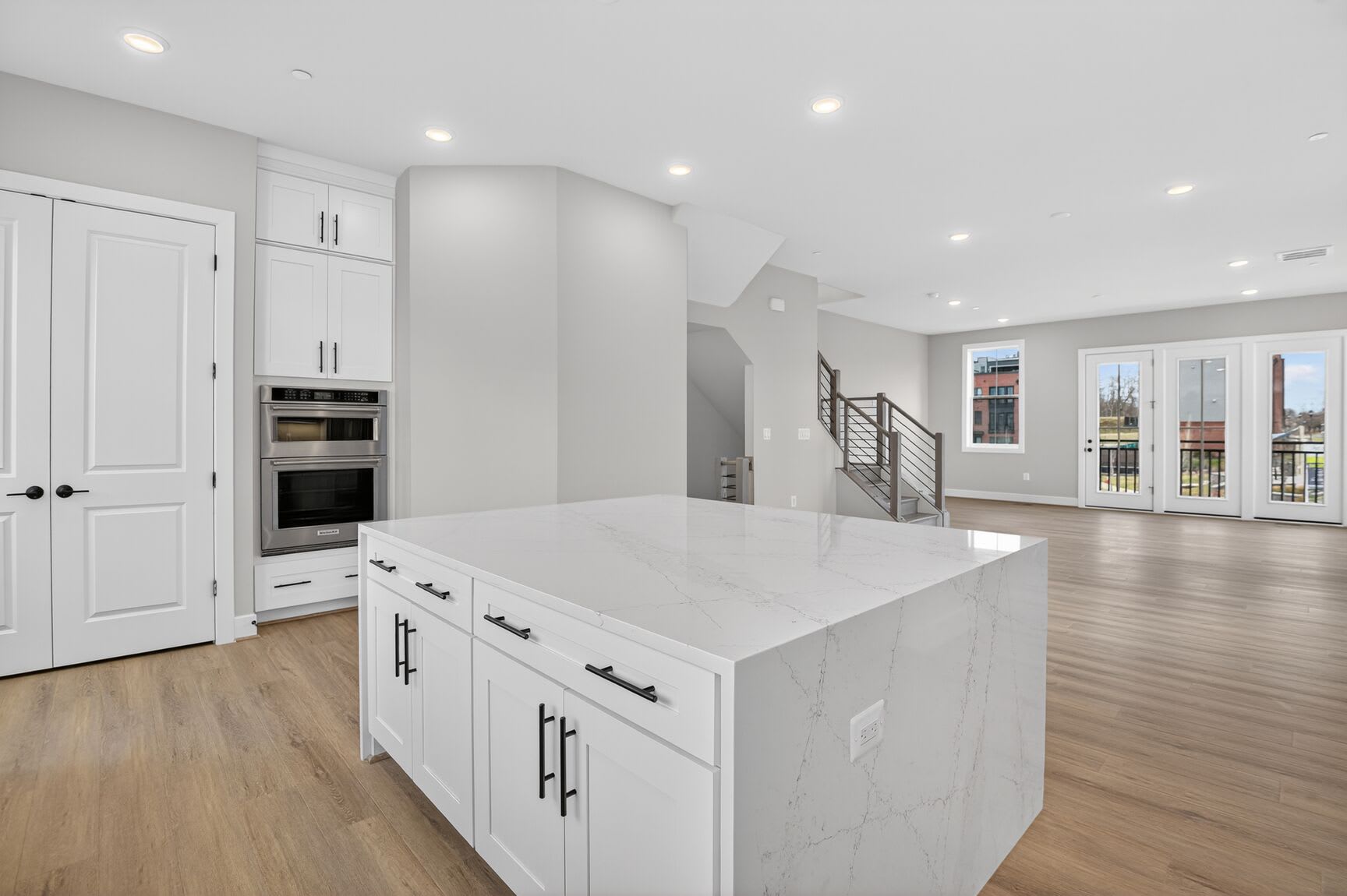 A modern, open-concept kitchen with white cabinets, a large island, and hardwood floors, with a staircase visible in the background.