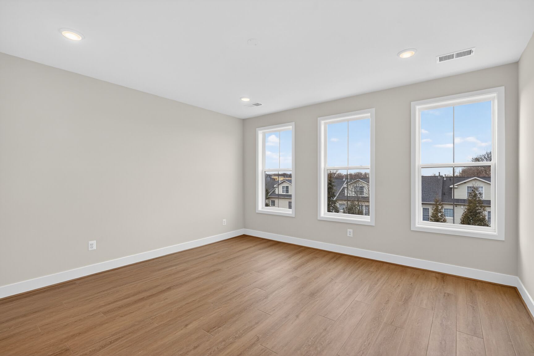 A spacious, well-lit room with large windows overlooking a residential neighborhood, featuring hardwood floors and neutral-colored walls.