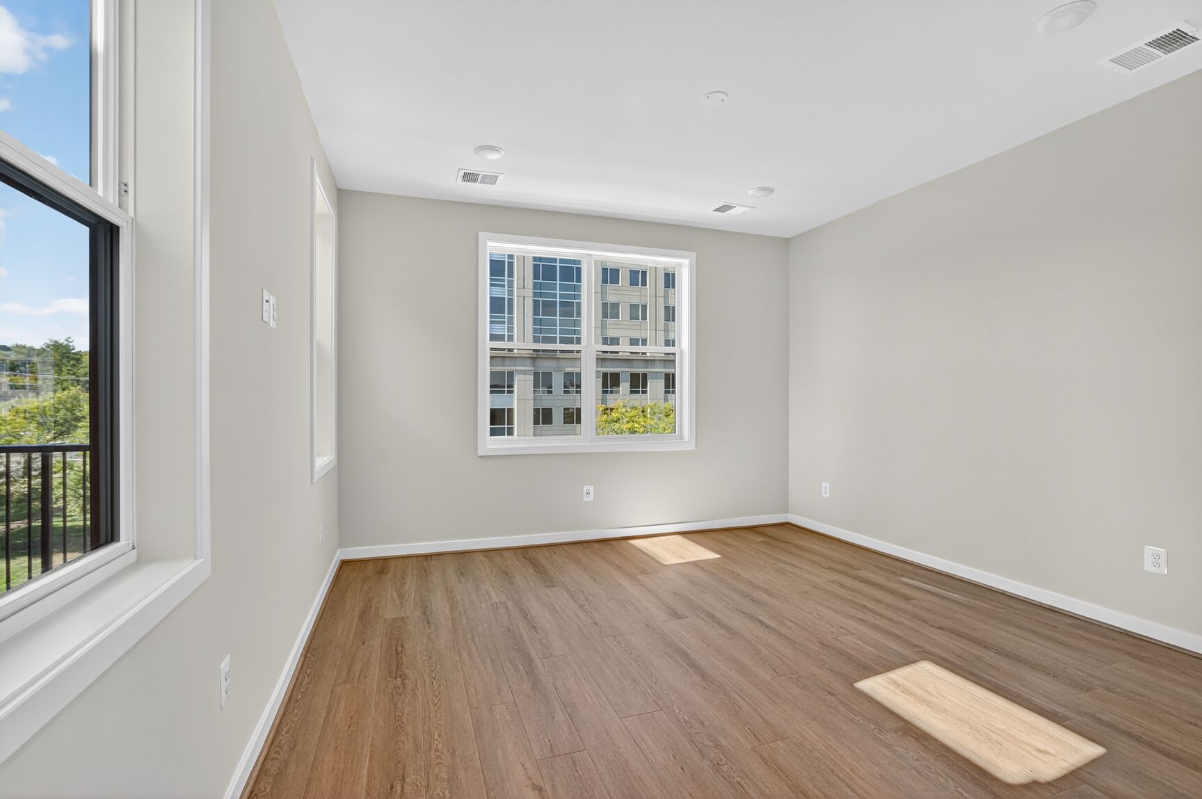 A spacious, well-lit room with a large window overlooking a scenic outdoor view, featuring hardwood floors and neutral-toned walls.