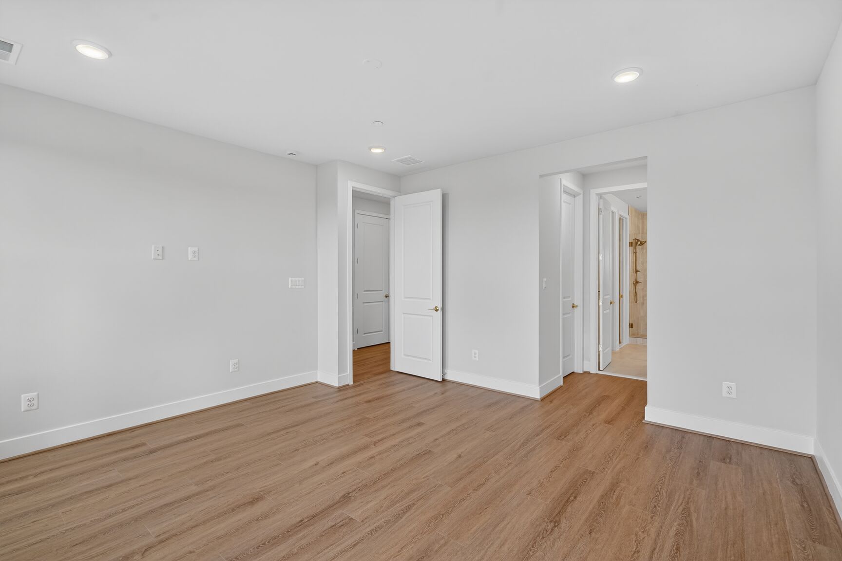 A spacious, well-lit room with white walls and hardwood floors, featuring several closed doors and recessed lighting.
