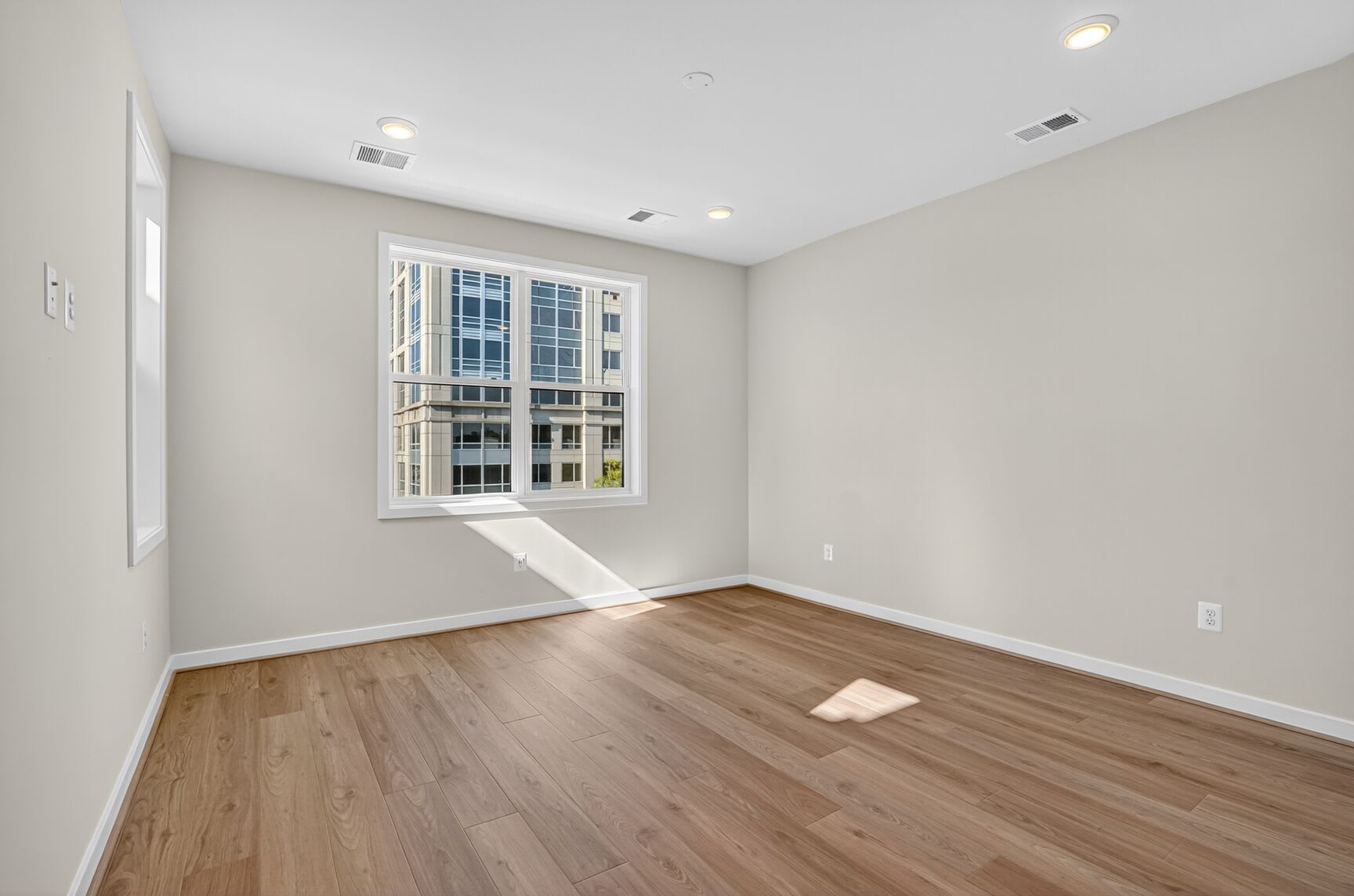 An empty room with hardwood floors, a large window, and recessed lighting in the ceiling.