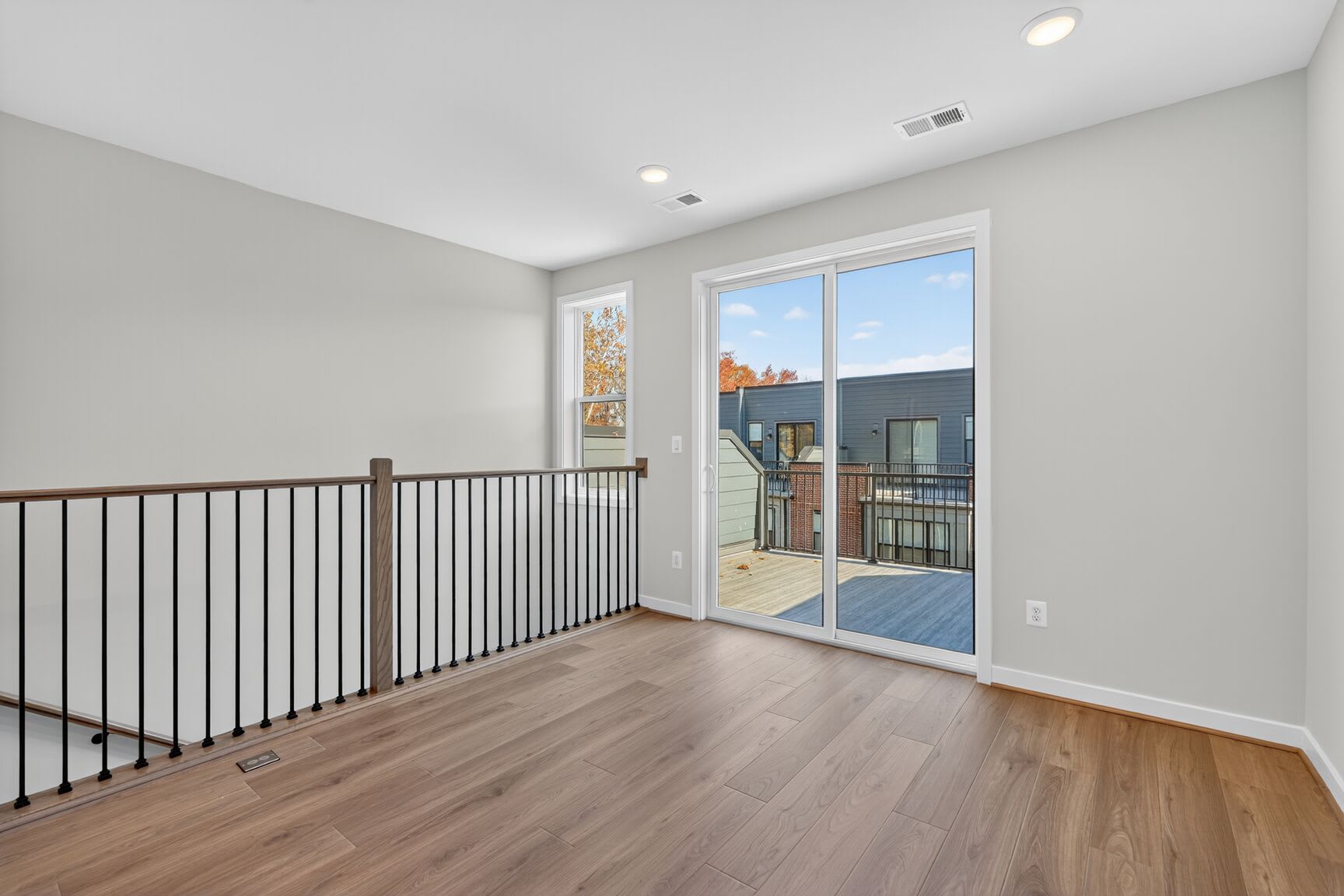A spacious room with hardwood floors, a glass sliding door leading to a balcony, and a metal railing along the upper level.