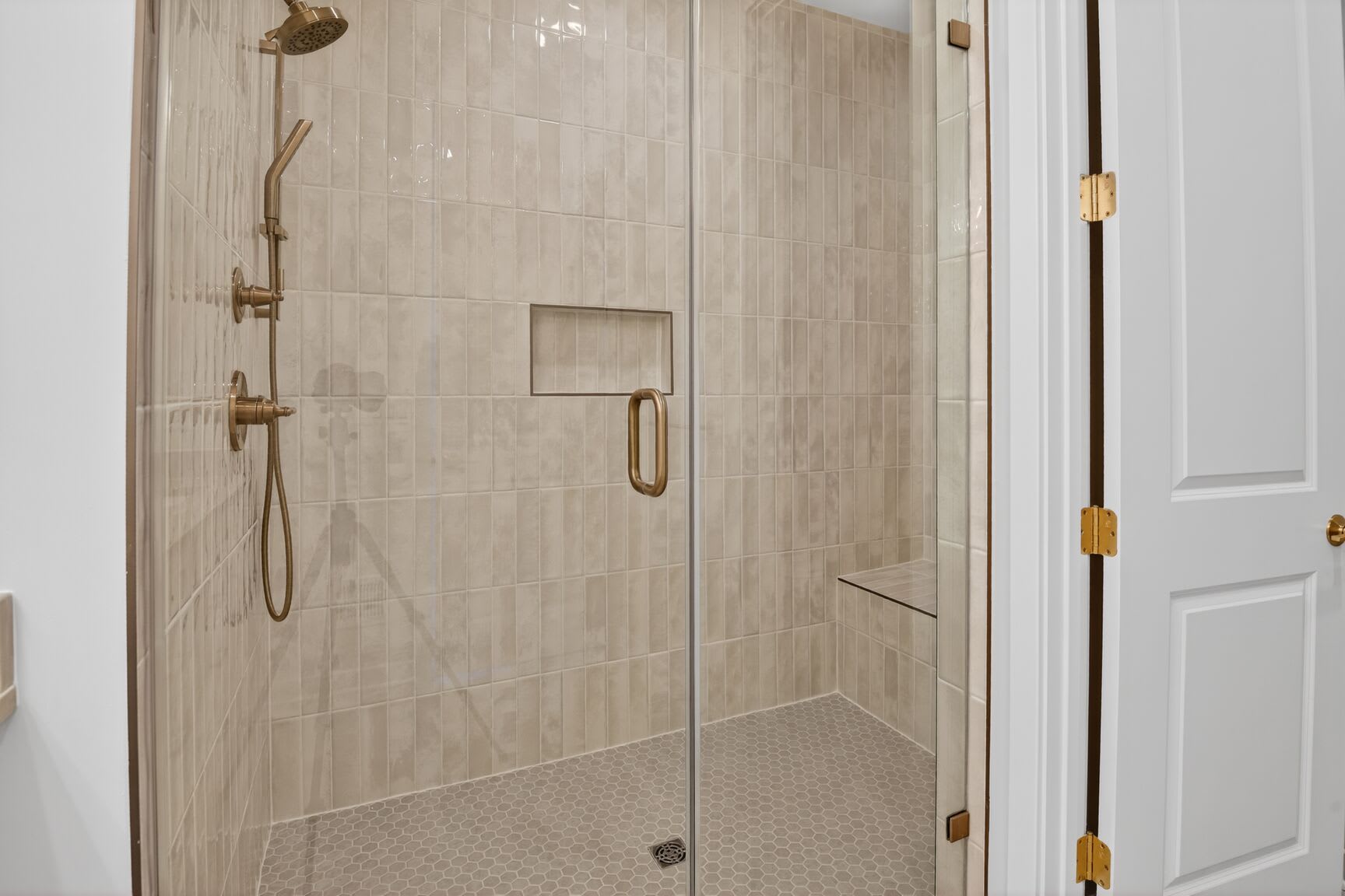 A modern, tiled shower enclosure with a glass door and brass fixtures, set against a white wall backdrop.