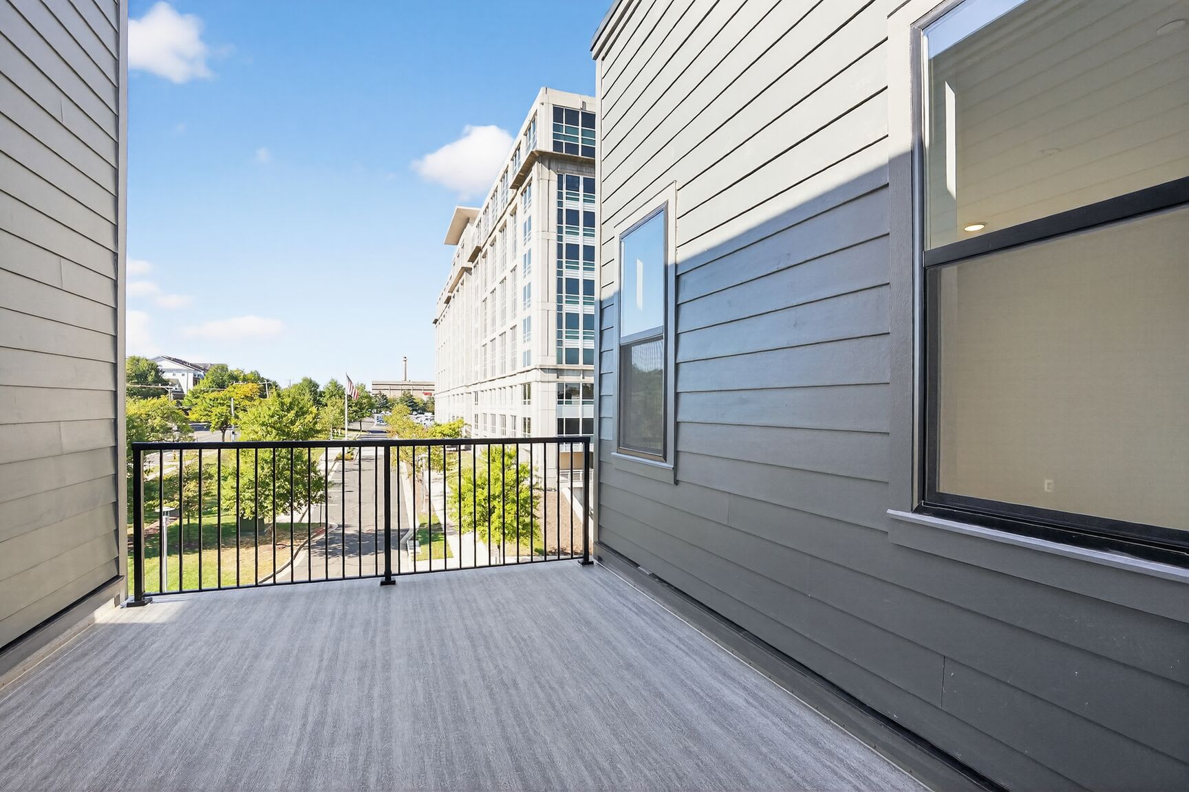A balcony with a wooden floor and metal railing, overlooking a modern apartment building with gray siding and large windows against a backdrop of a blue sky with scattered clouds.