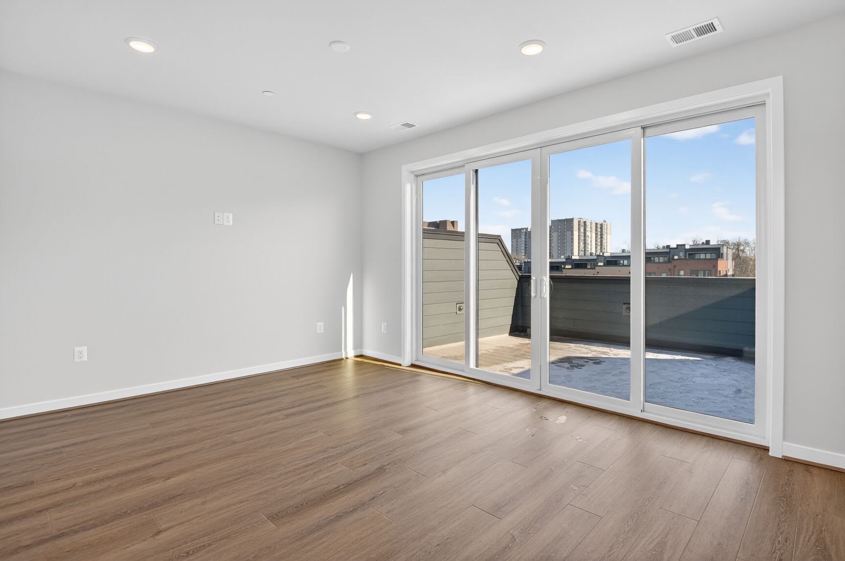 A bright, spacious room with large windows overlooking a cityscape, featuring hardwood floors and a sliding glass door leading to a balcony or terrace.
