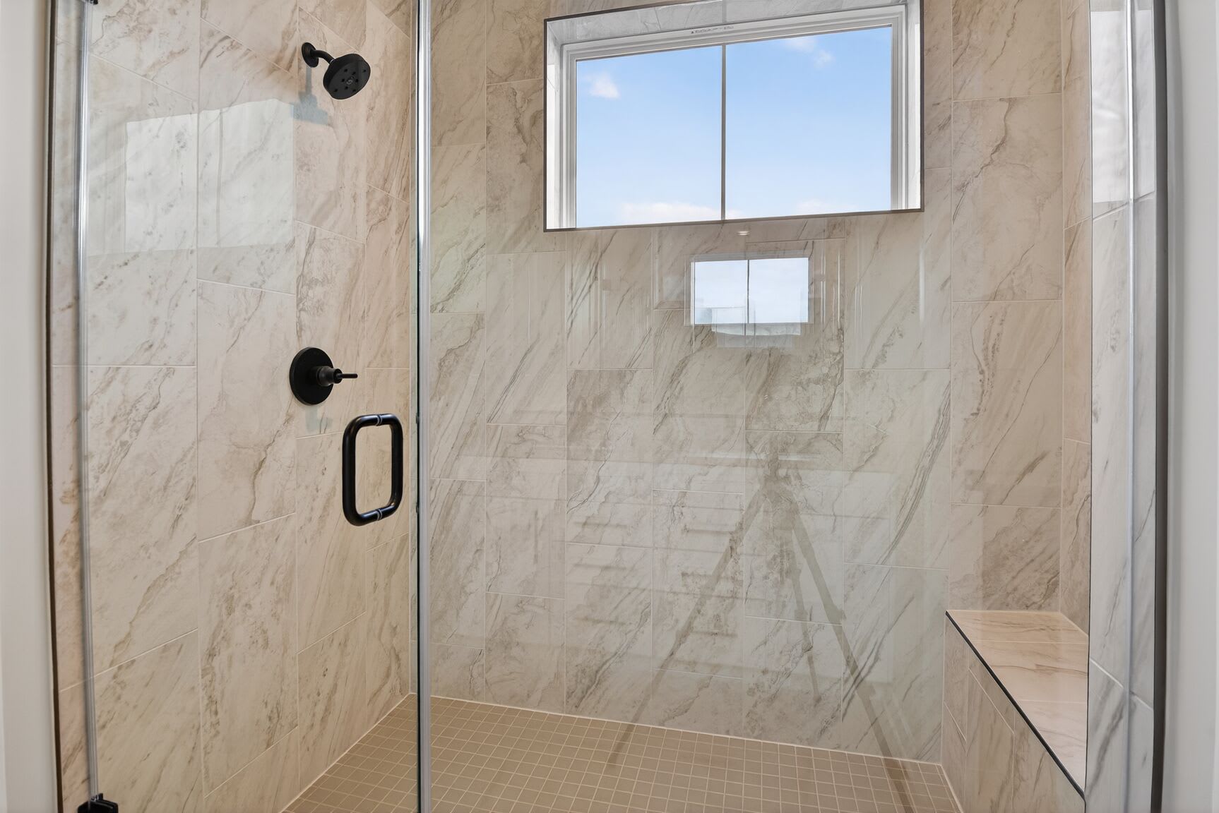 A modern, minimalist bathroom with a glass shower enclosure, beige marble-like tiles on the walls, and a small window providing natural light.