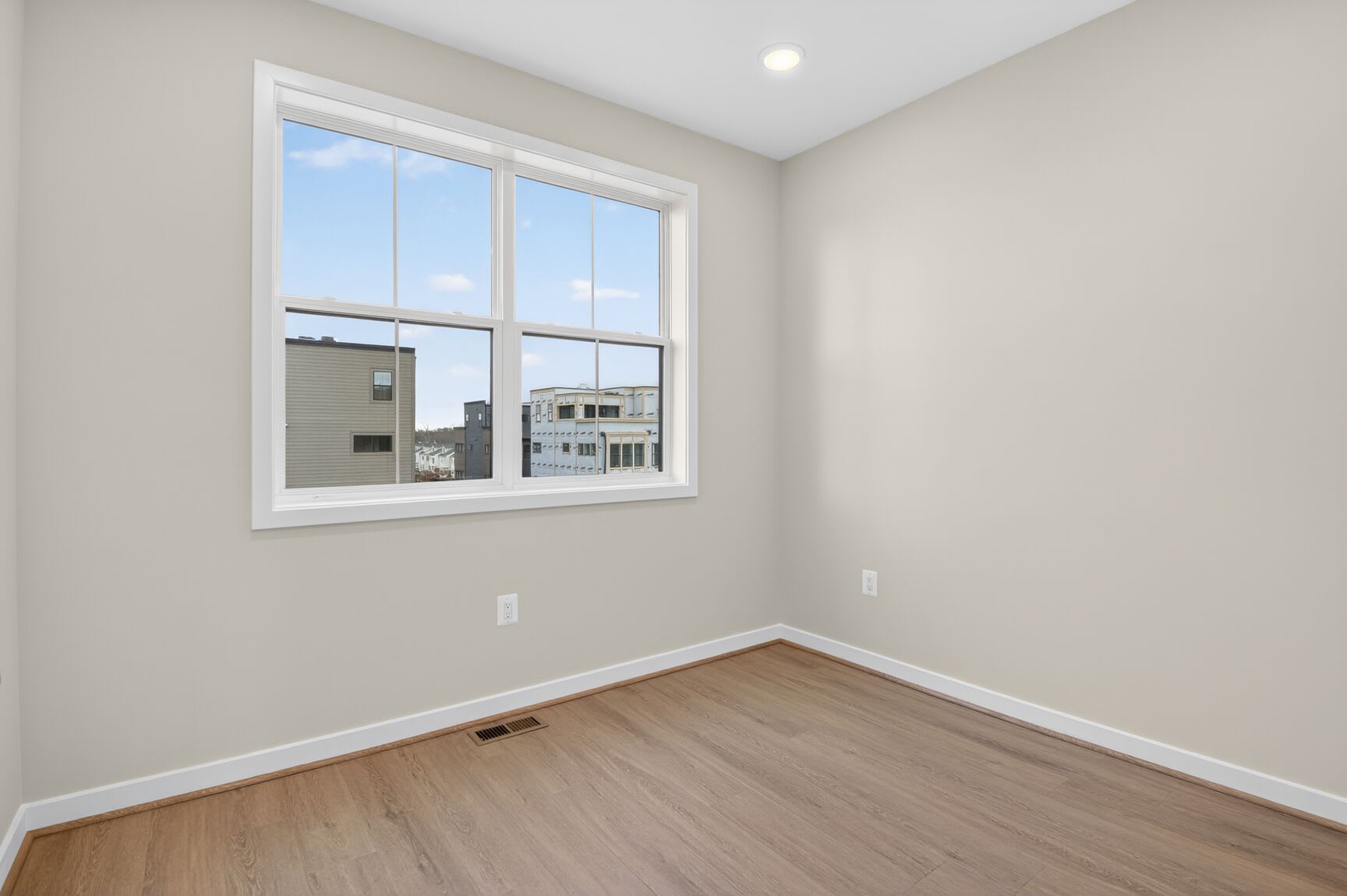 A spacious, well-lit room with a large window overlooking a cityscape, featuring a hardwood floor and neutral-colored walls.