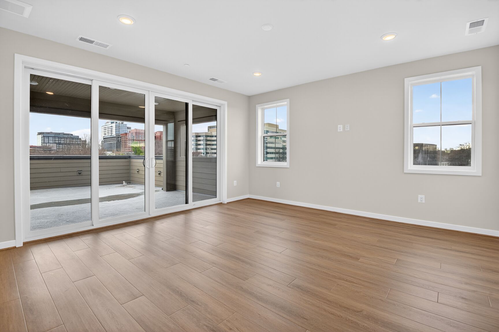 A spacious, well-lit room with hardwood floors, large windows, and sliding glass doors leading to a balcony or patio, offering a view of the surrounding cityscape.