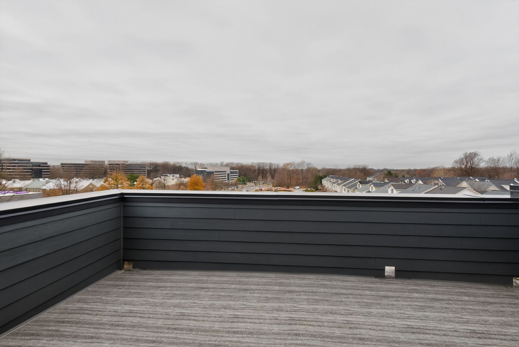 A wooden deck with a black railing overlooks a cityscape in the distance, with buildings and trees visible against a cloudy sky.