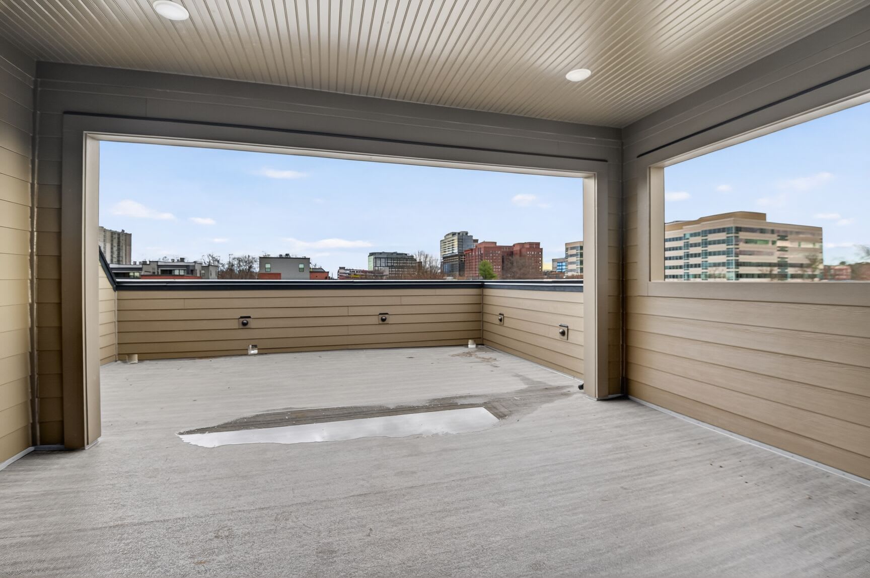 A spacious balcony or patio with a view of a city skyline, featuring a wooden floor, gray walls, and recessed lighting.