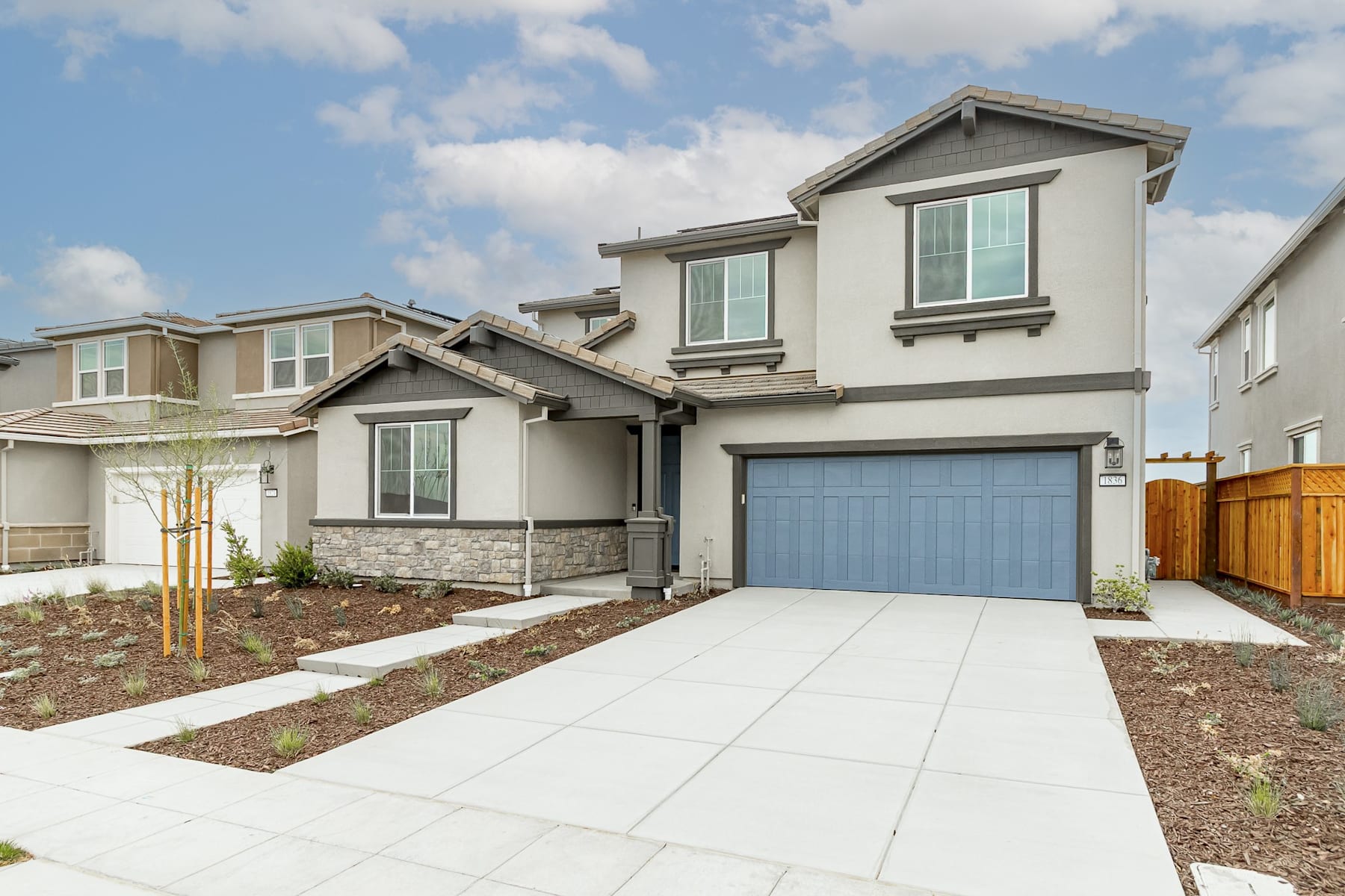 A modern two-story residential house with a garage, surrounded by a landscaped yard and a clear blue sky with scattered clouds in the background.