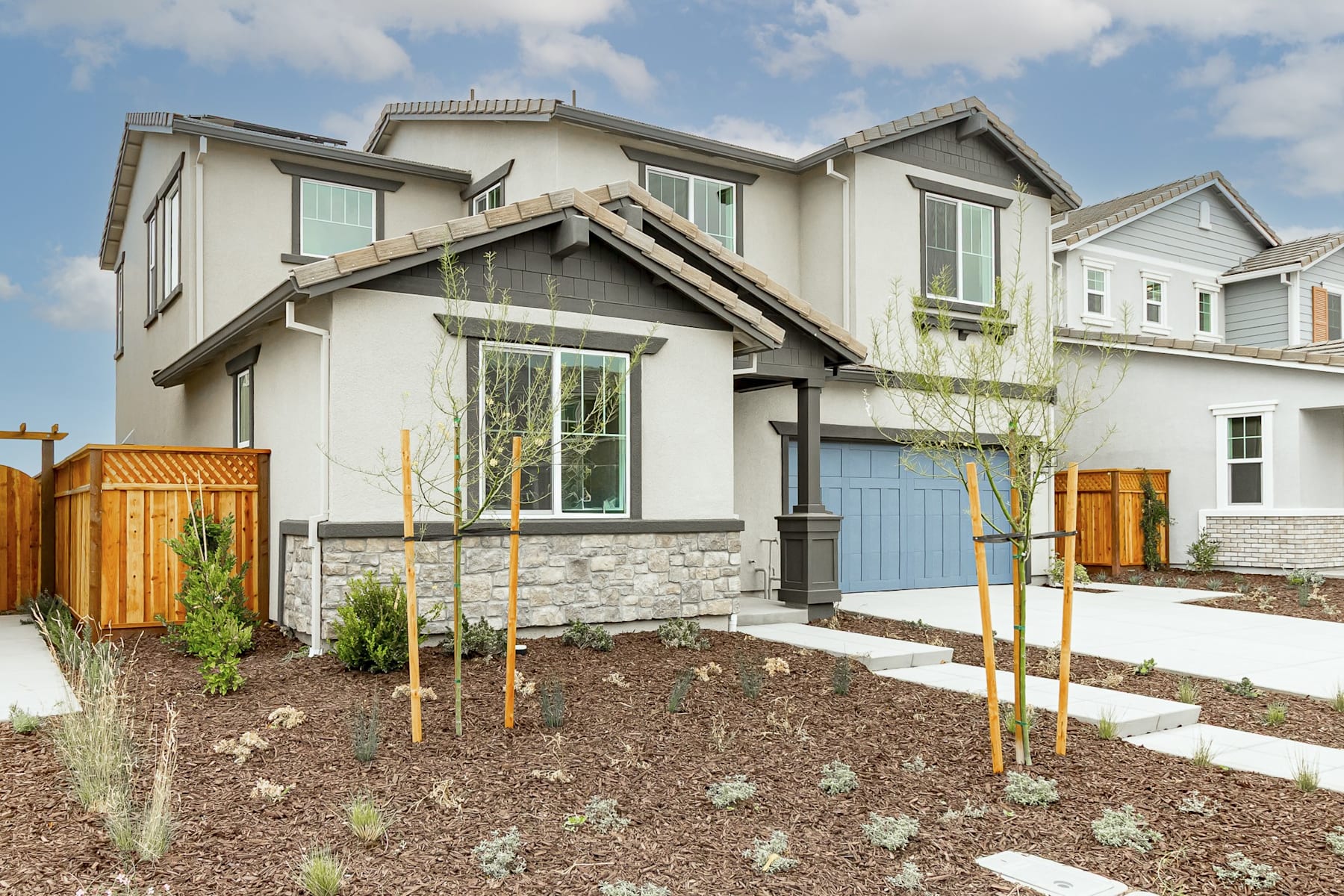 A modern two-story residential home with a landscaped front yard featuring newly planted trees and shrubs, set against a backdrop of a partly cloudy sky.