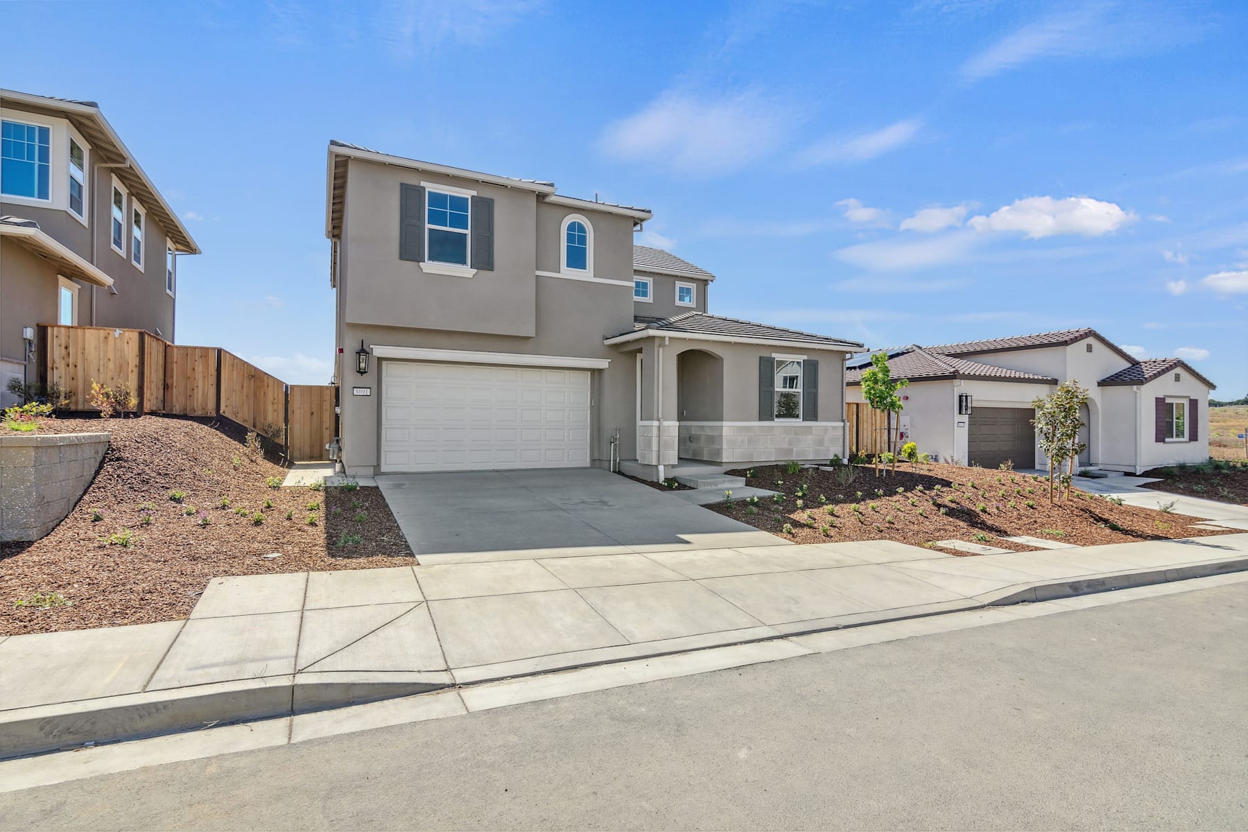 A two-story residential house with a garage, surrounded by a paved driveway and landscaped yard, set against a clear blue sky with scattered clouds.