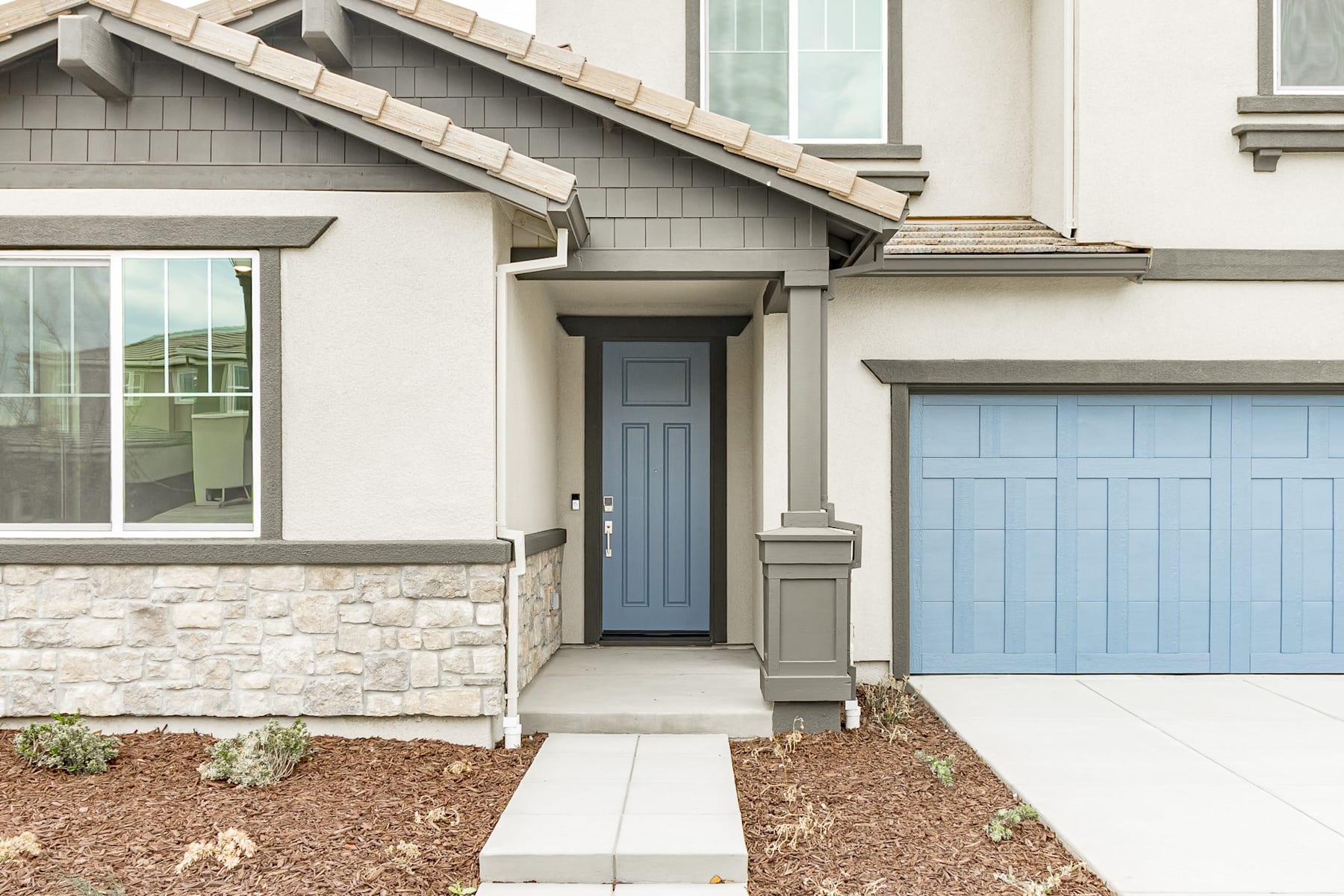 A modern, two-story house with a stone facade, a blue front door, and a matching blue garage door, surrounded by a landscaped yard with mulch and plants.