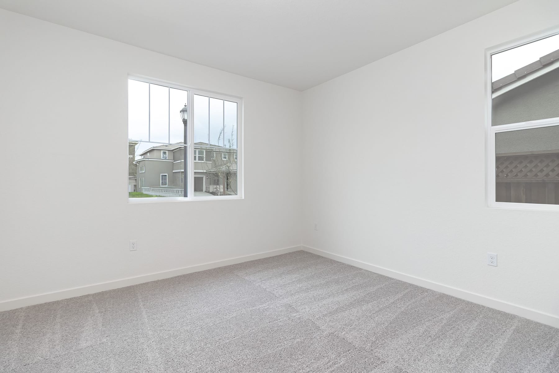 A bright, empty room with white walls, a hardwood floor, and large windows overlooking a residential neighborhood in the background.