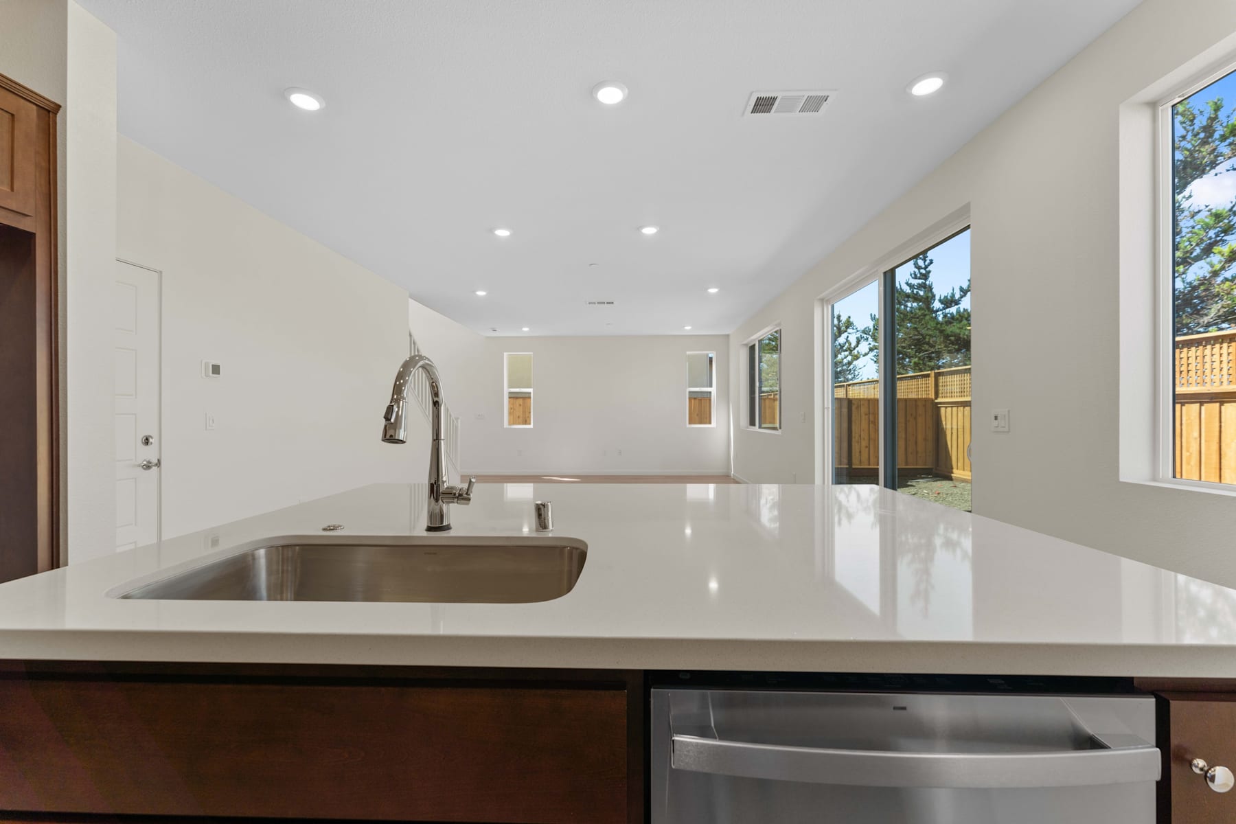 A modern, open-concept kitchen with a large stainless steel sink, sleek countertops, and recessed lighting, set against a backdrop of bright, airy windows overlooking a fenced outdoor area.