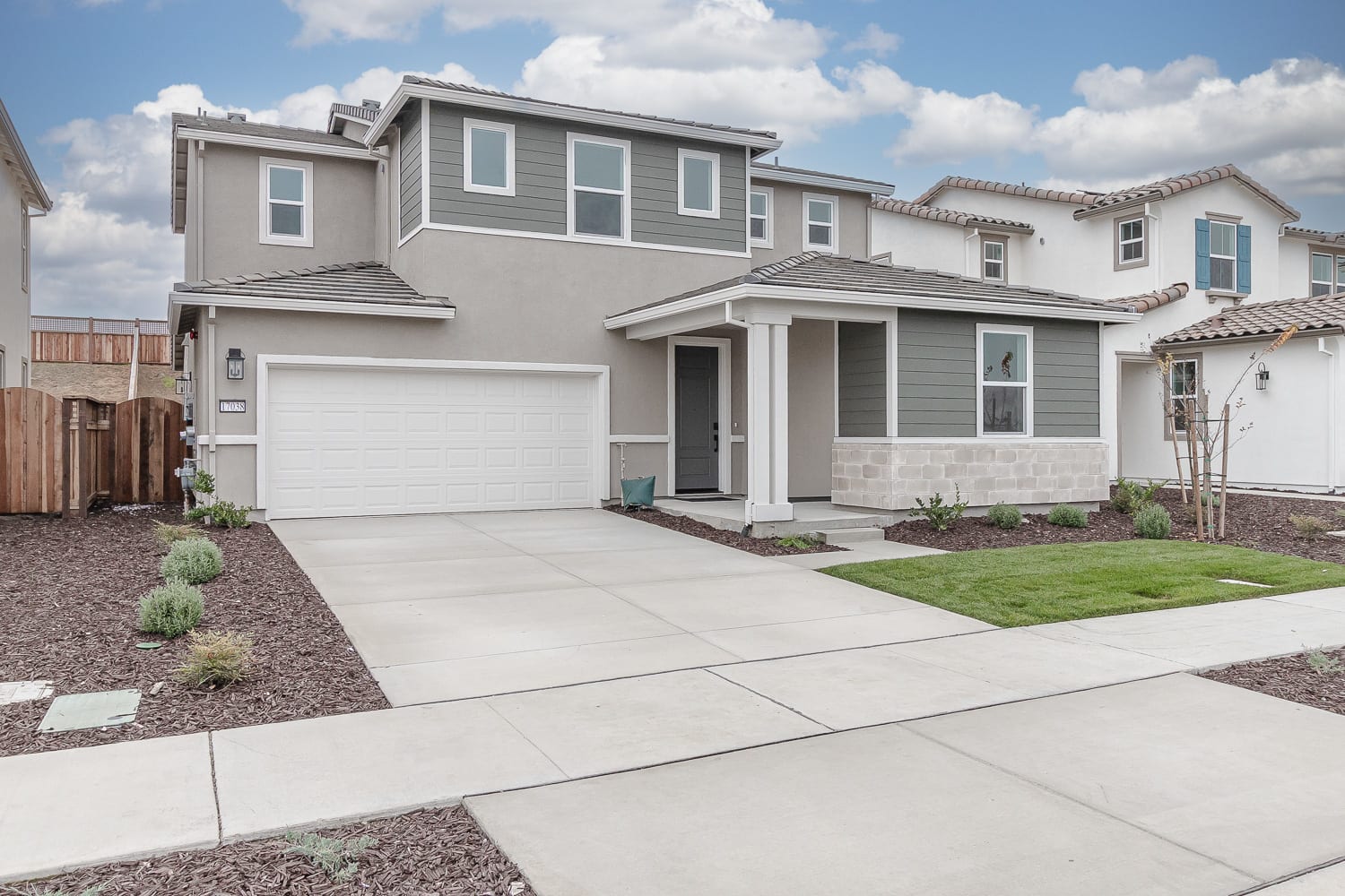 A two-story residential house with a garage, surrounded by a paved driveway and a well-manicured lawn, set against a backdrop of a partly cloudy sky.