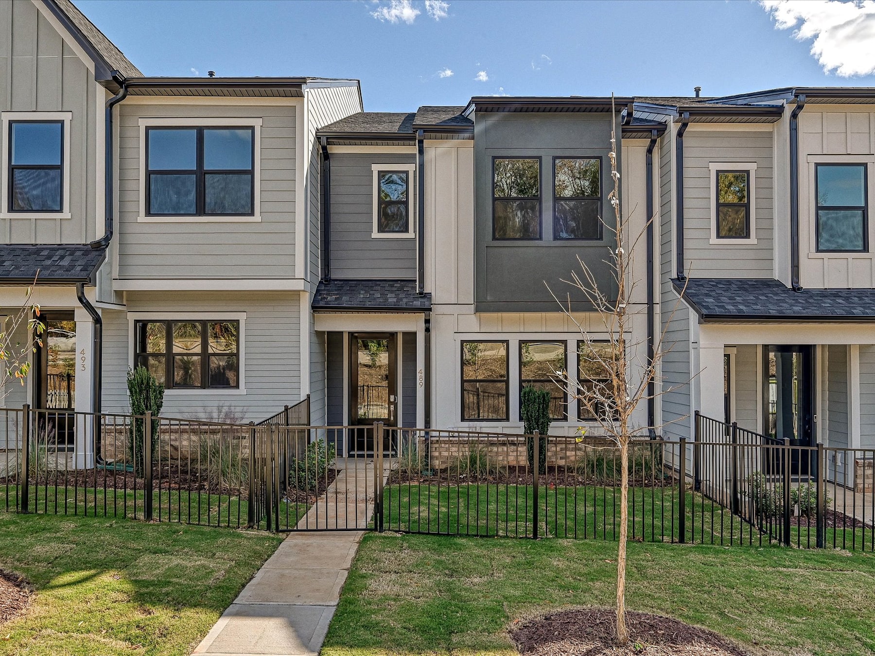 A row of modern, multi-story townhouses with gray and beige siding, surrounded by a grassy lawn and a metal fence in the foreground.