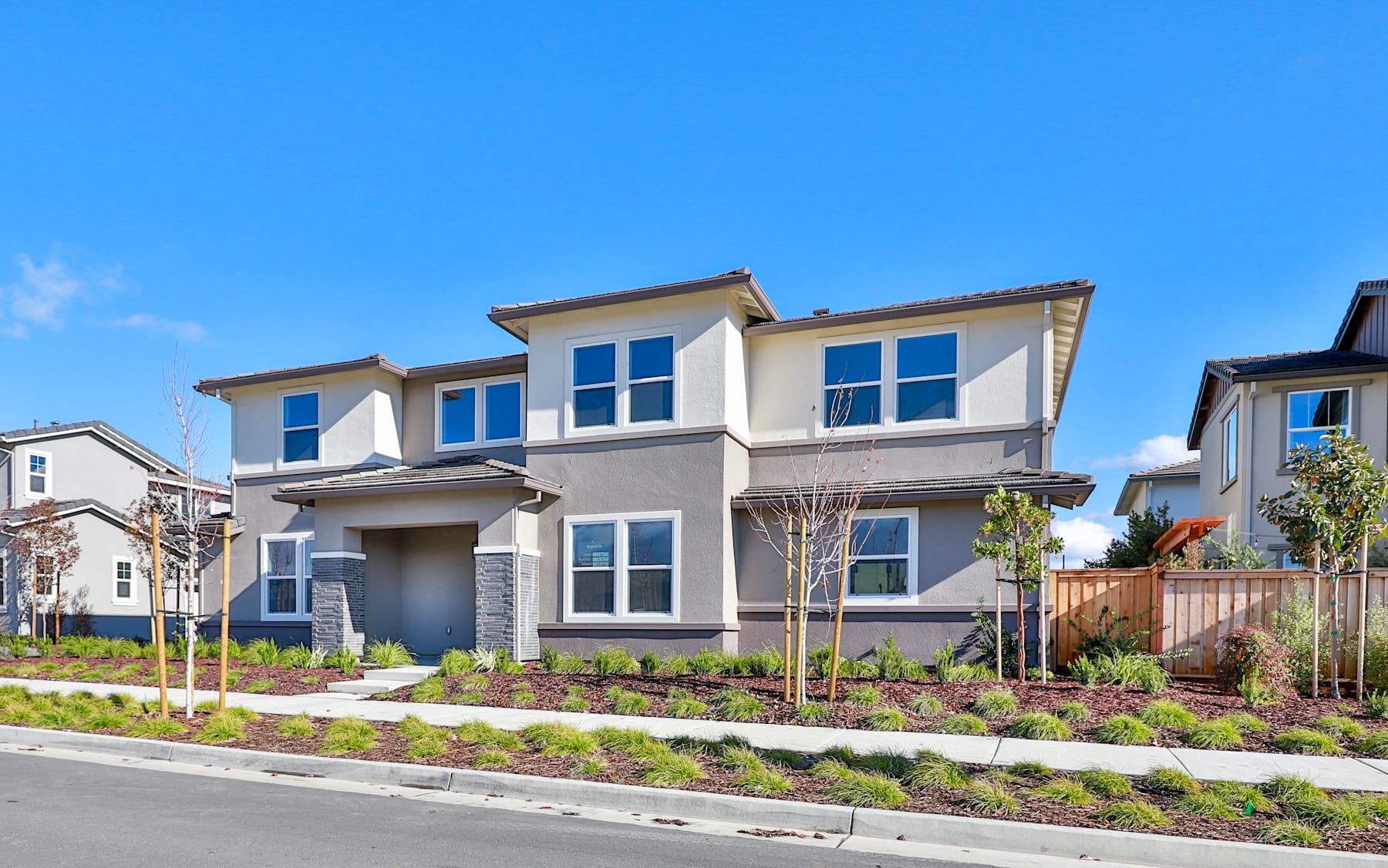 A modern, two-story residential building with a well-landscaped front yard, set against a clear blue sky.