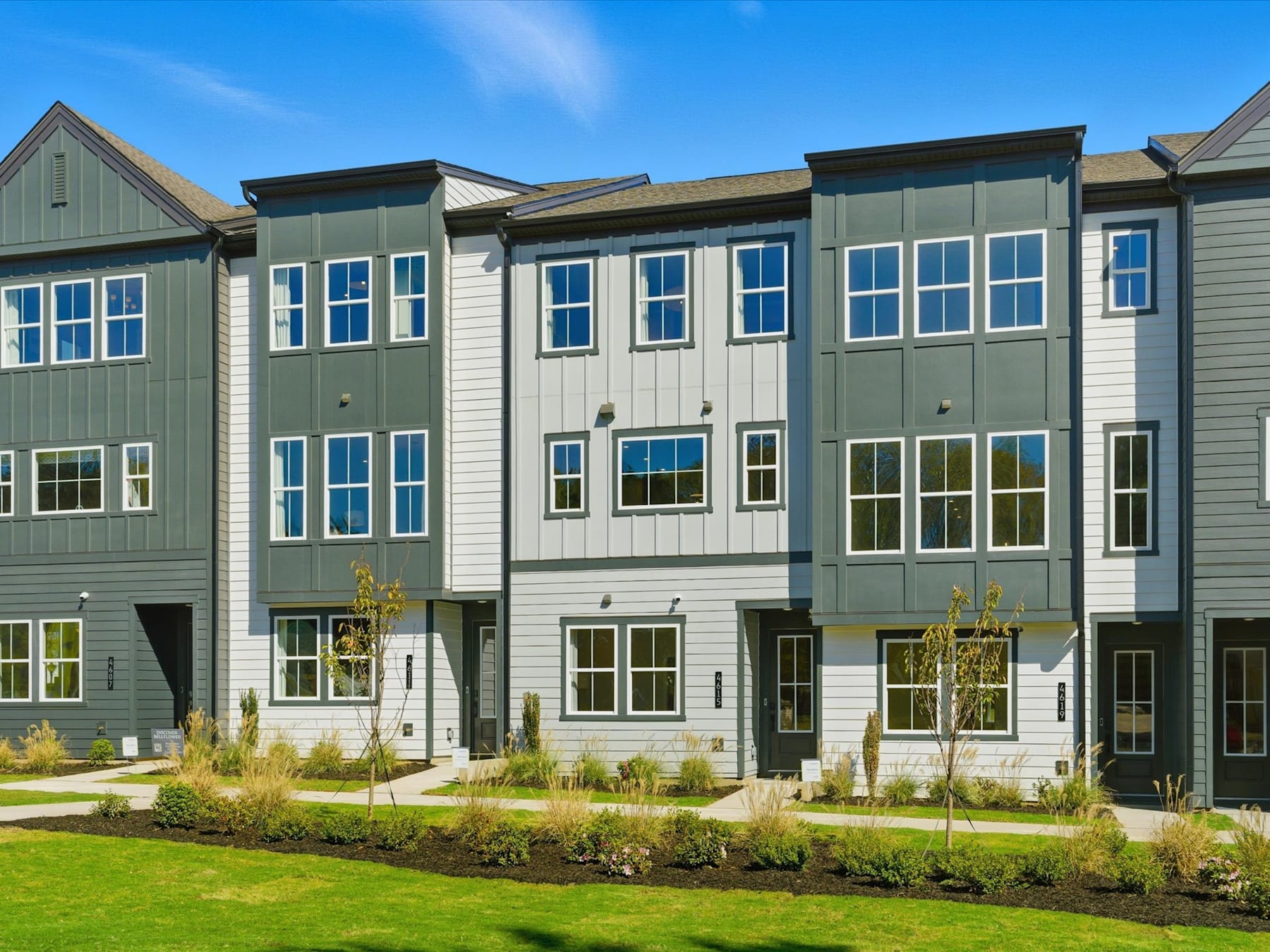A row of modern, multi-story townhouses with green and gray siding, surrounded by a well-manicured lawn and landscaping in the foreground, set against a clear blue sky in the background.