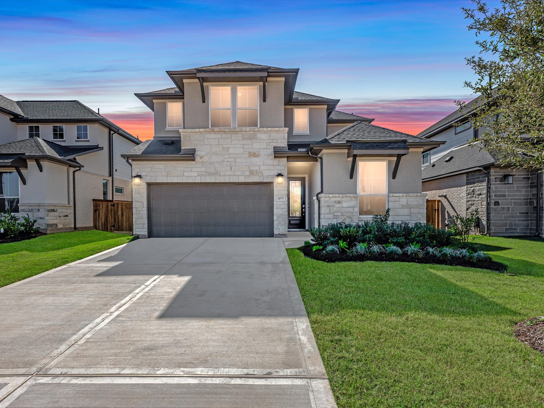 A modern, two-story house with a stone facade stands in a well-manicured yard, surrounded by a clear sky and a vibrant sunset in the background.
