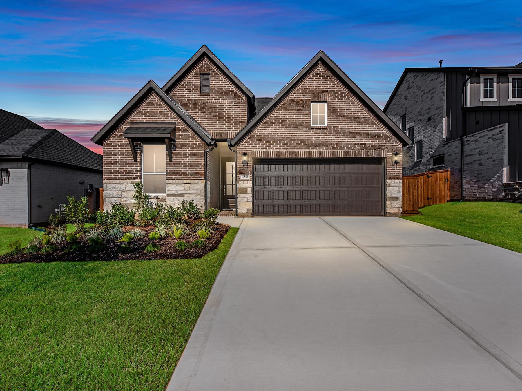 A modern two-story brick house with a garage and a well-manicured lawn in the foreground, set against a vibrant sky with a colorful sunset in the background.
