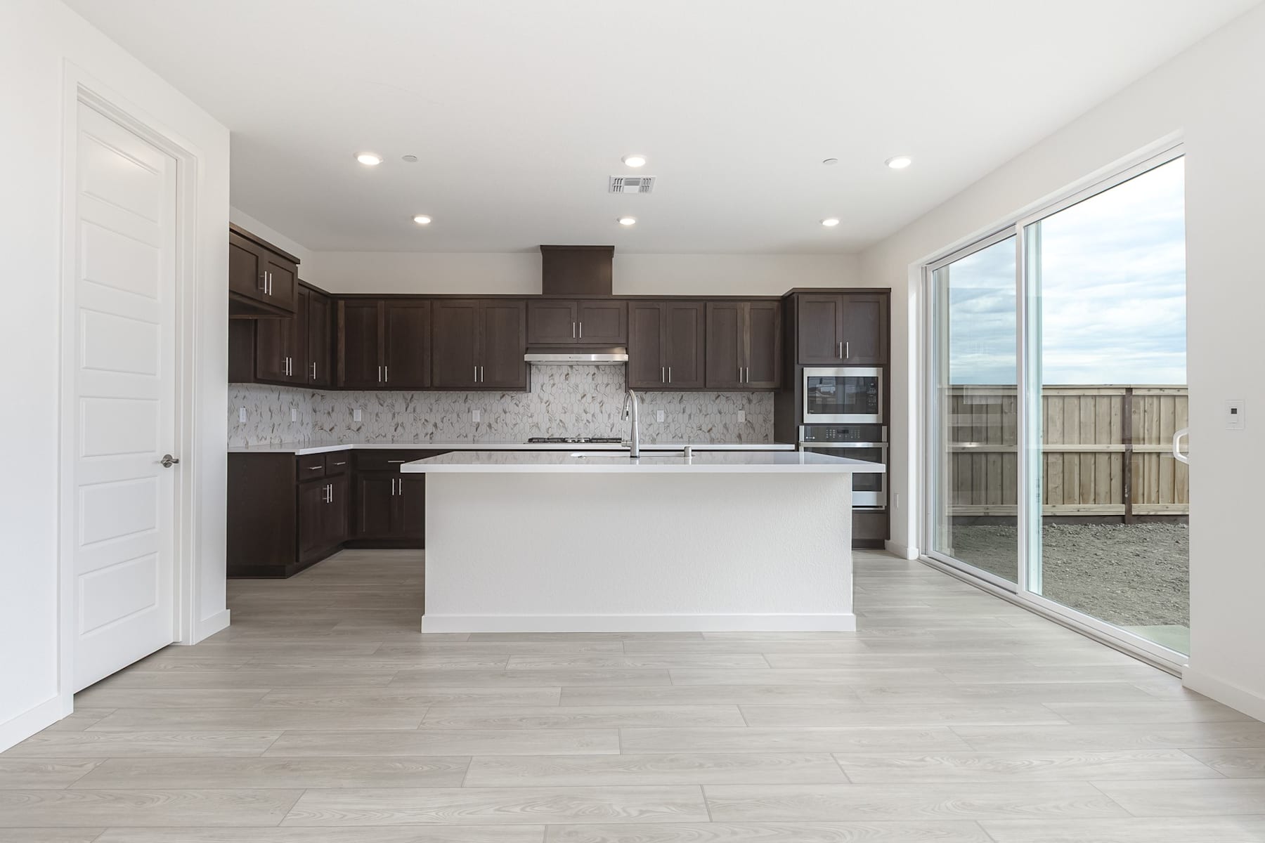 A modern, open-concept kitchen with dark wood cabinets, a white island, and large windows overlooking an outdoor area.