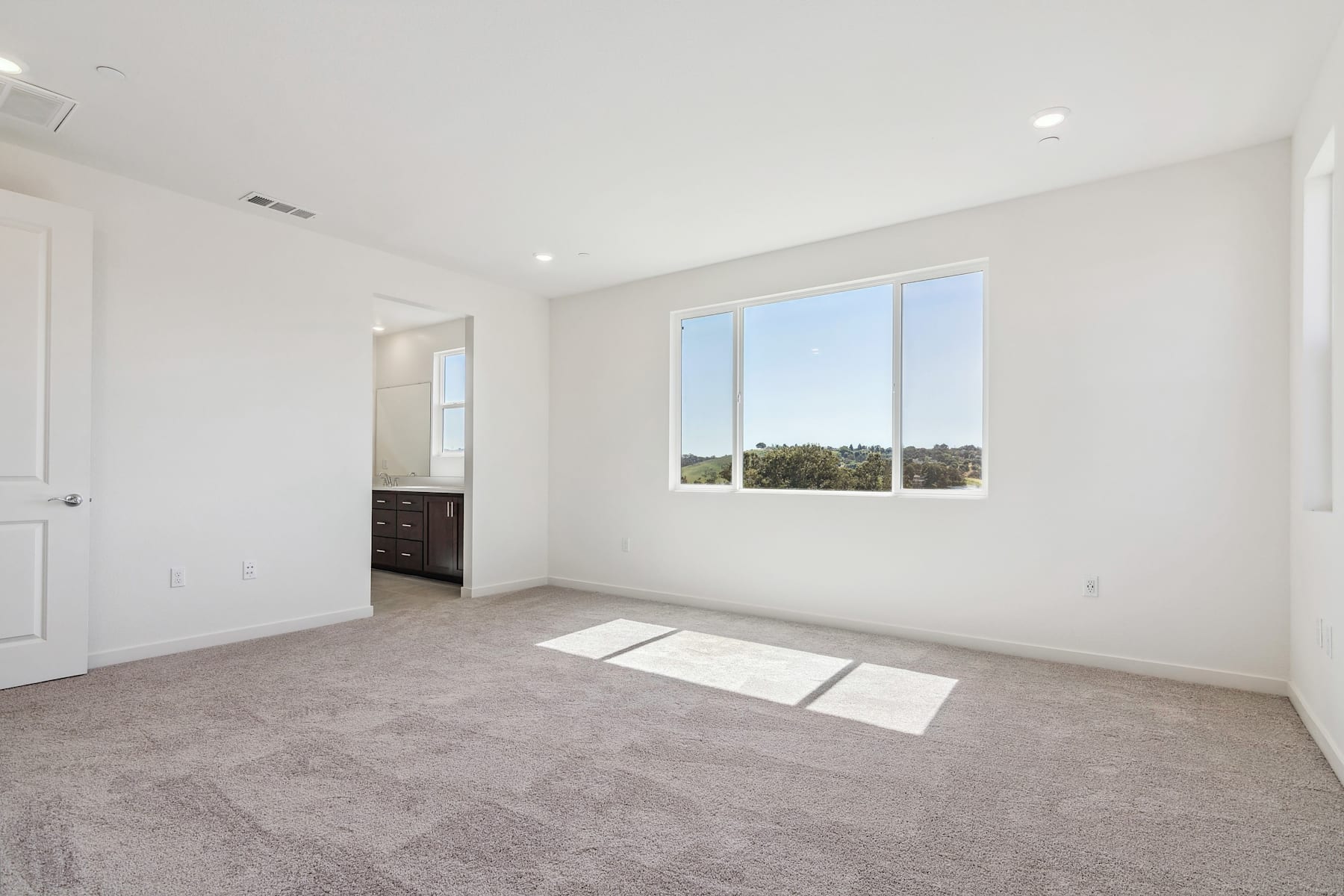 A spacious, well-lit bedroom with a large window overlooking a scenic outdoor view, featuring a carpeted floor and a door leading to an adjacent room.