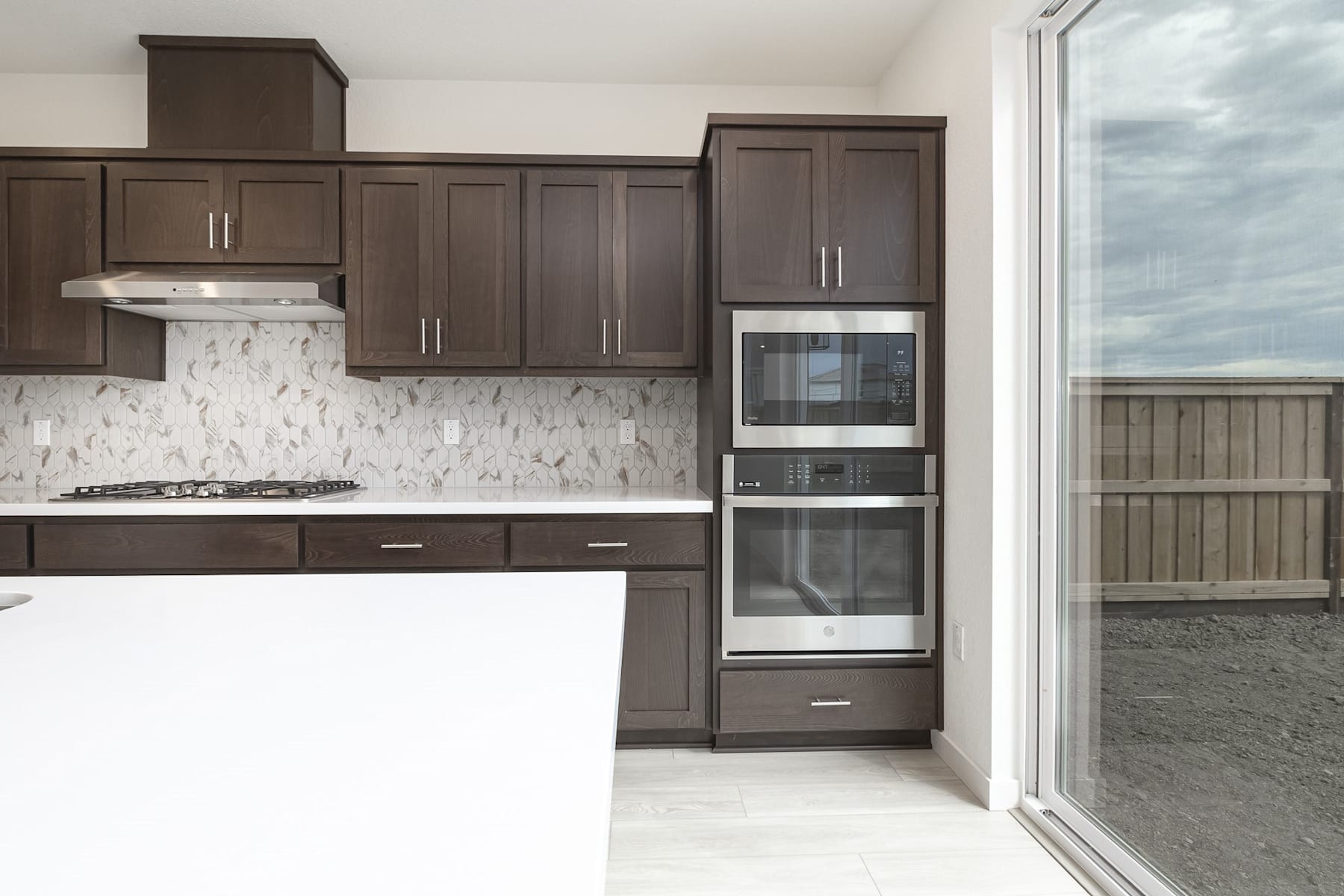 A modern kitchen with dark wood cabinets, a white marble backsplash, and stainless steel appliances, set against a backdrop of a sliding glass door leading to an outdoor area.