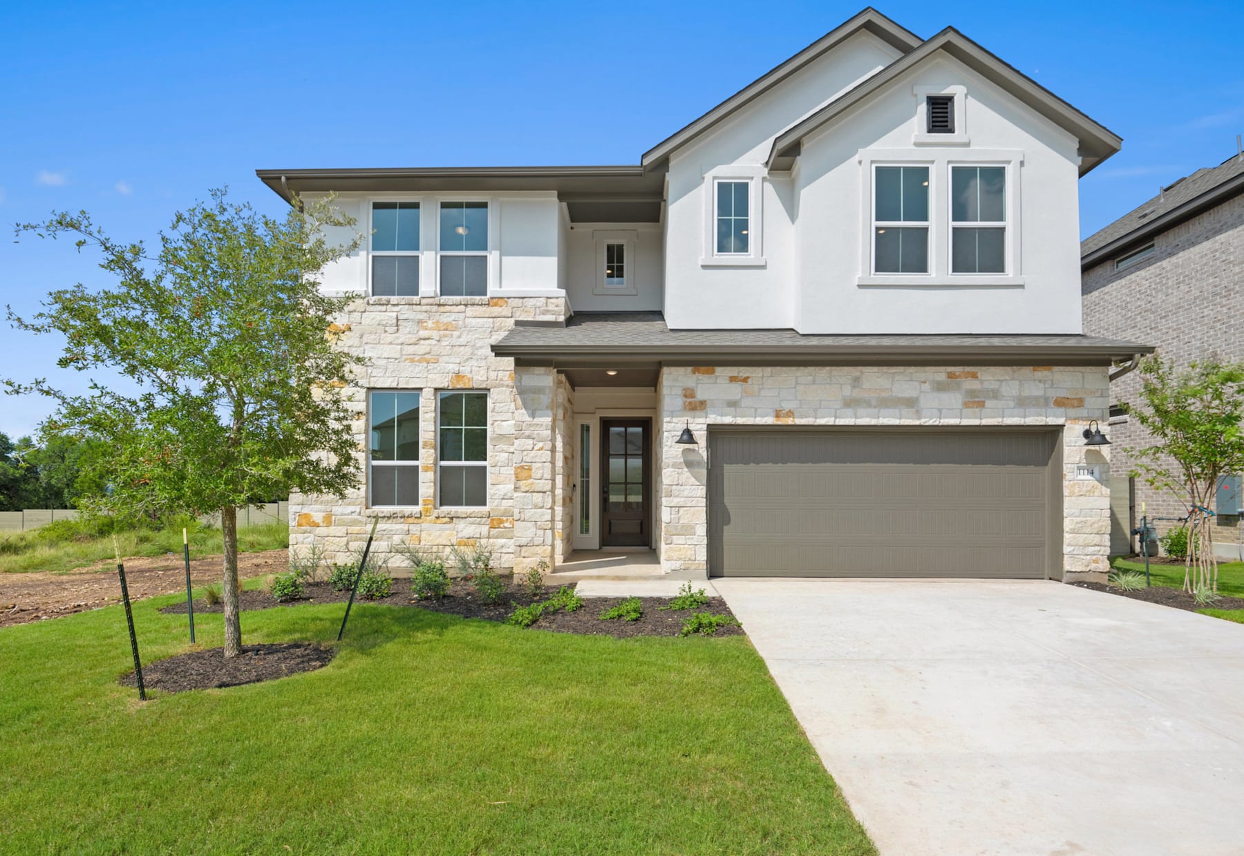 A two-story modern house with a stone exterior, a garage, and a well-manicured lawn surrounded by lush greenery against a clear blue sky.