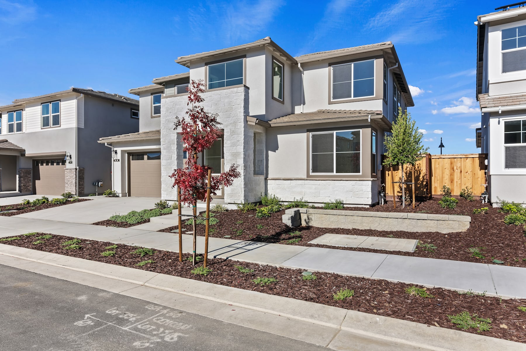 A modern, two-story residential building with a well-landscaped front yard, featuring a paved walkway, shrubs, and a decorative tree in the foreground, set against a clear blue sky.