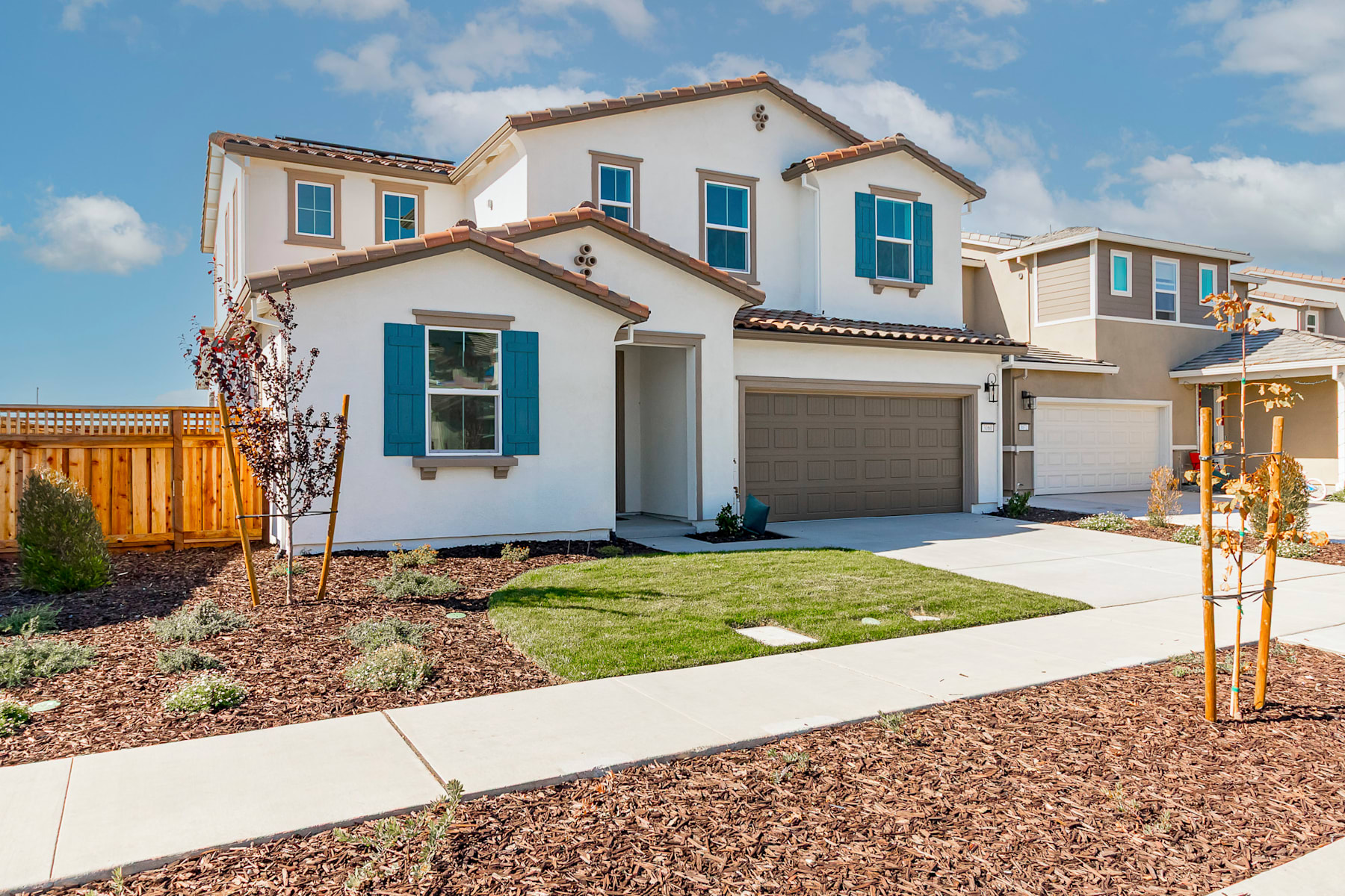 A two-story residential house with a garage, surrounded by a landscaped yard with a concrete walkway leading to the front entrance, set against a backdrop of a blue sky with scattered clouds.